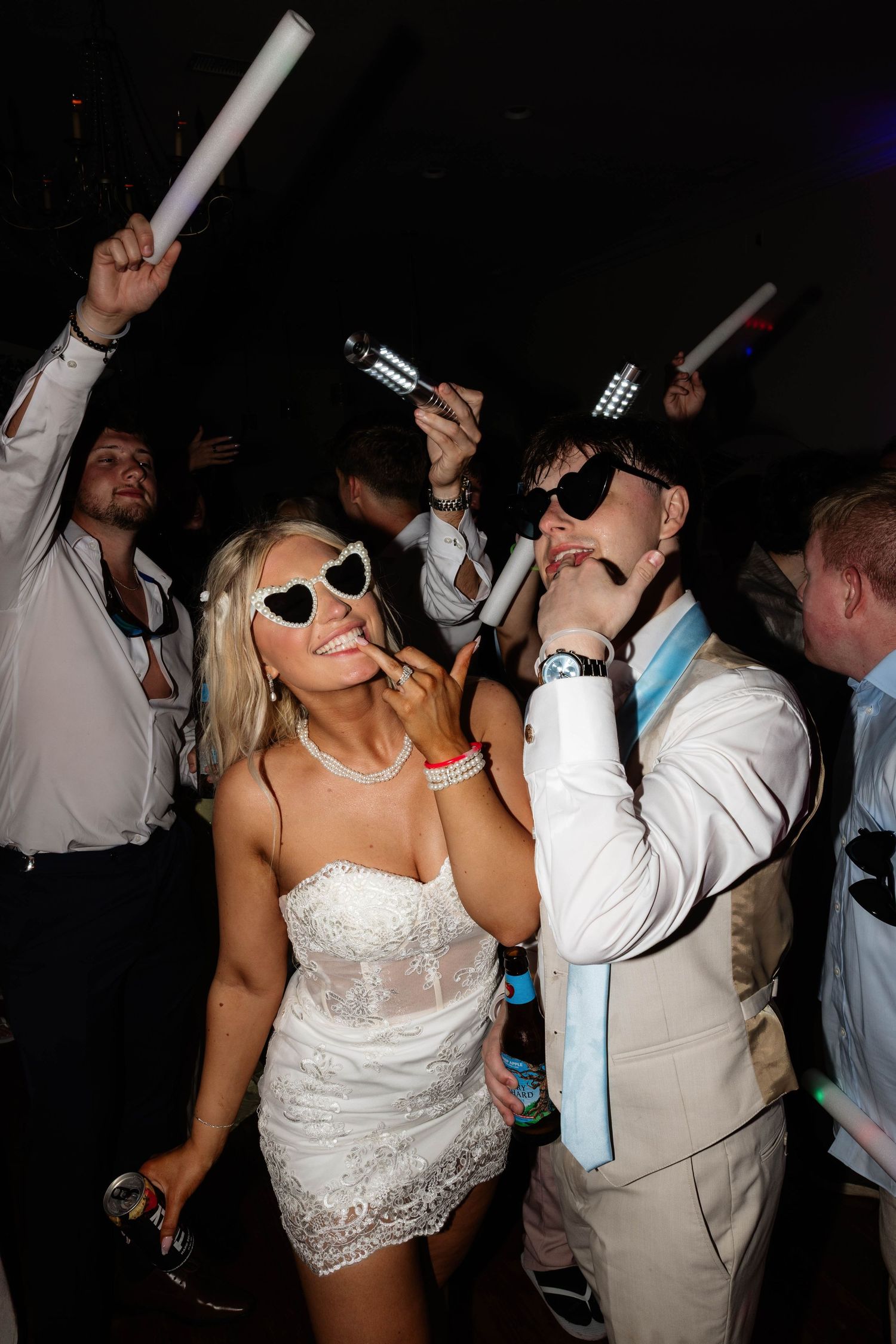 Bride in white mini dress and heart sunglasses dancing with groom in white suit at lively wedding reception.