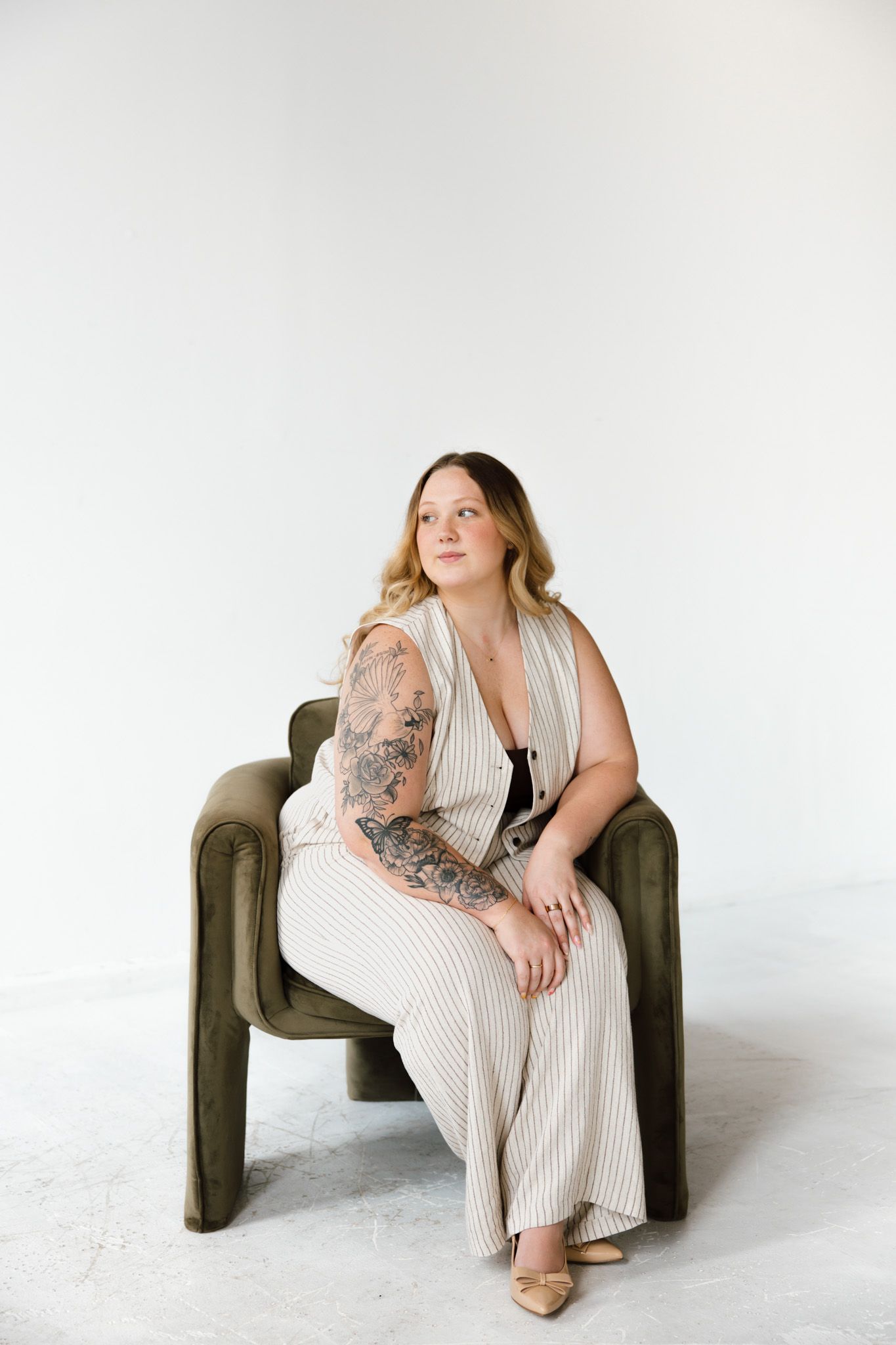 Woman with tattoos in striped jumpsuit sitting in olive green chair against white background.