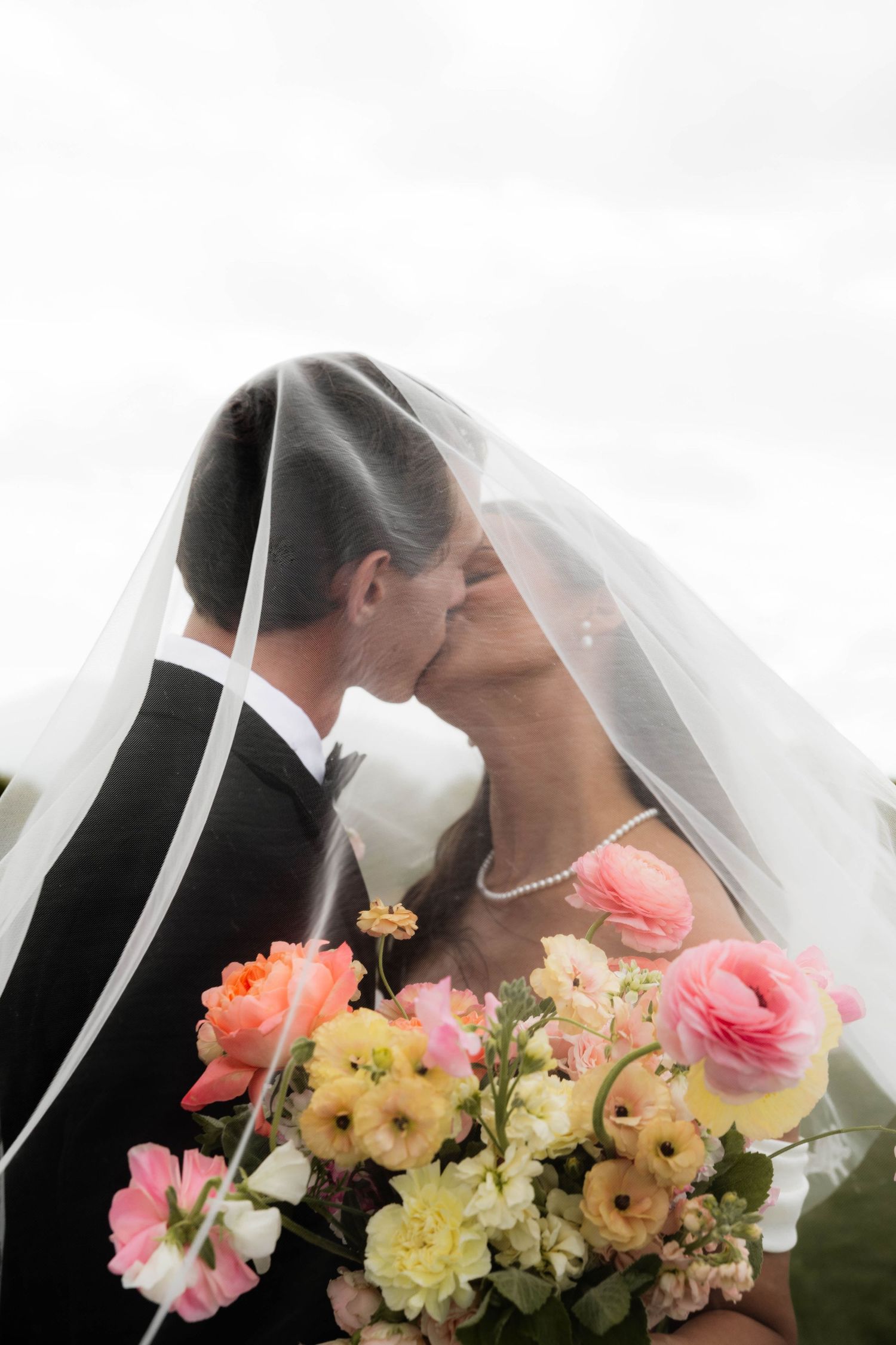Bride and groom kissing under a wedding veil, holding a colorful bouquet of pink and yellow flowers.