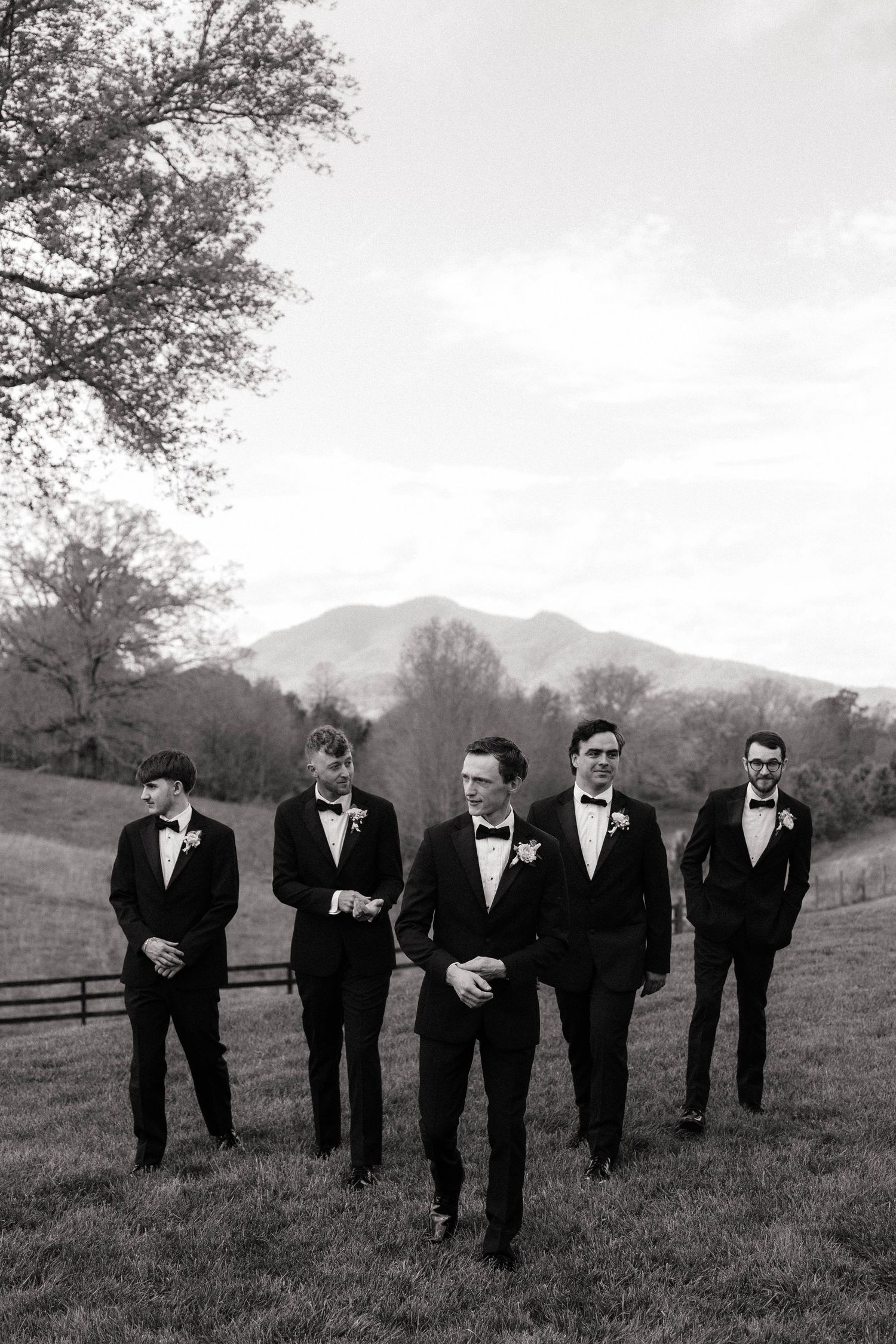 Black and white photo of groom and groomsmen in tuxedos walking outdoors with mountain backdrop.