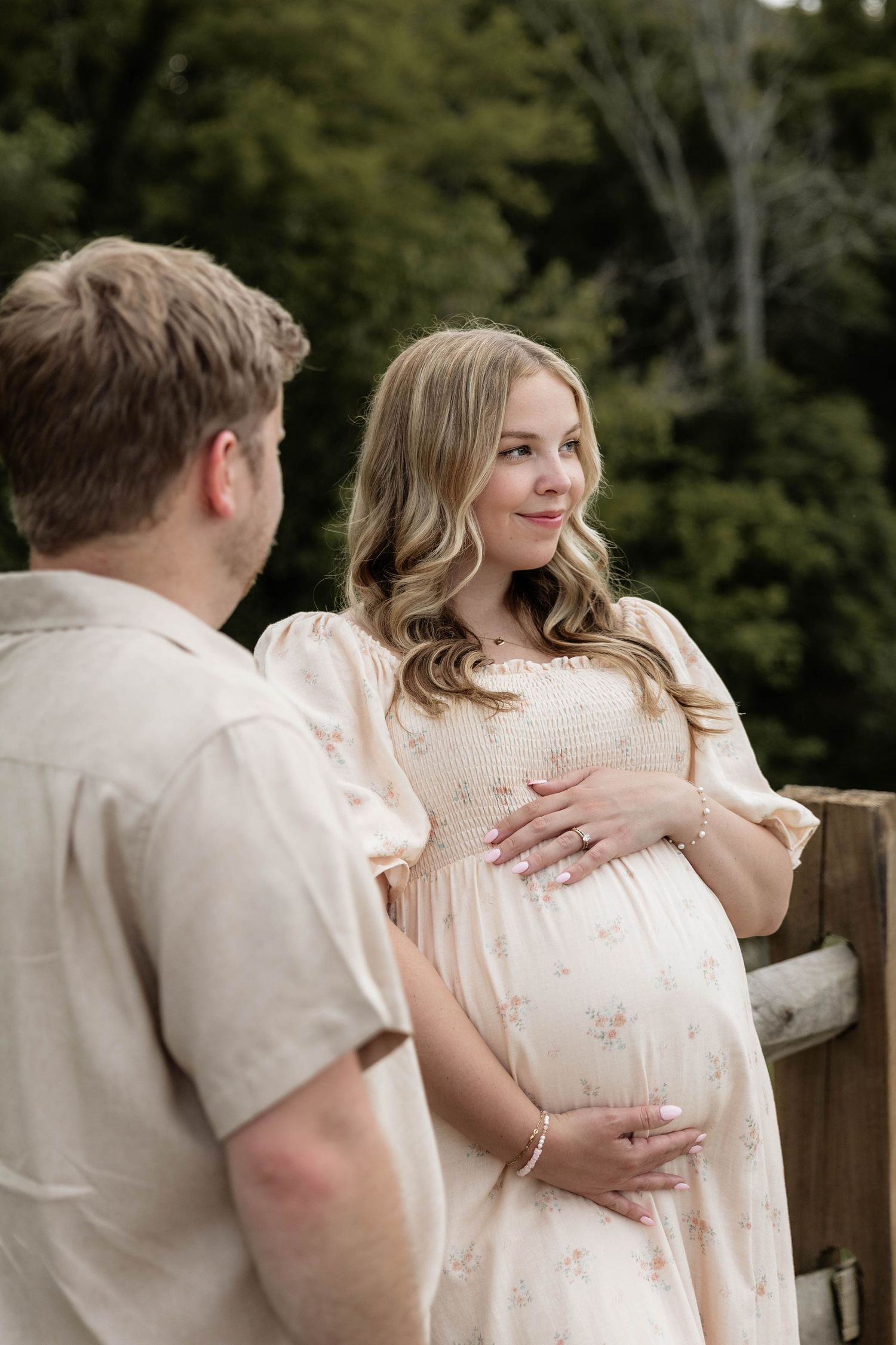 Pregnant woman in floral dress cradling baby bump while smiling at partner outdoors near wooden fence.