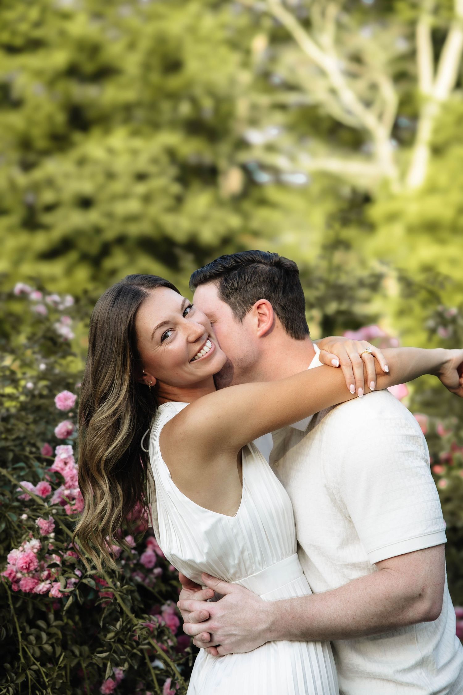Couple embracing in a garden with pink flowers, woman in white dress smiling, man hugging her from behind.