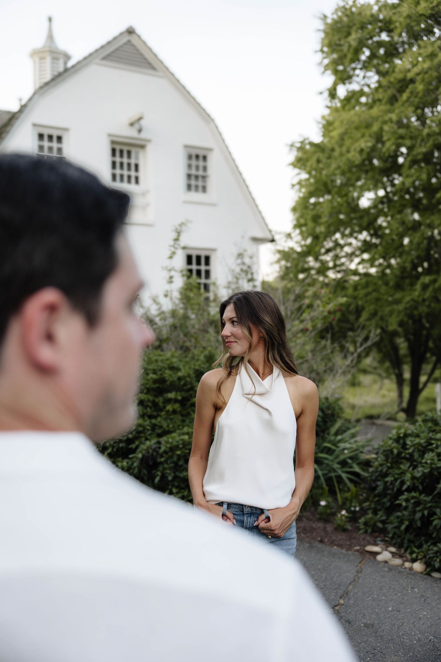 Woman in white halter top and jeans smiling outdoors near a white house with lush greenery.