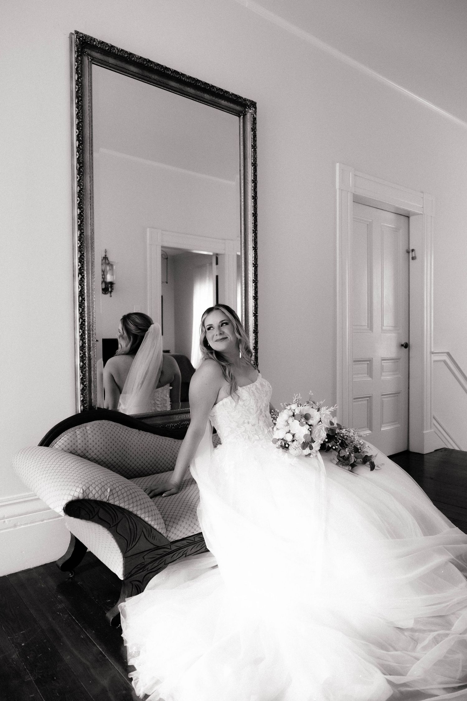 Bride in a white tulle gown holding a bouquet sits before a large ornate mirror in a black and white bridal portrait.