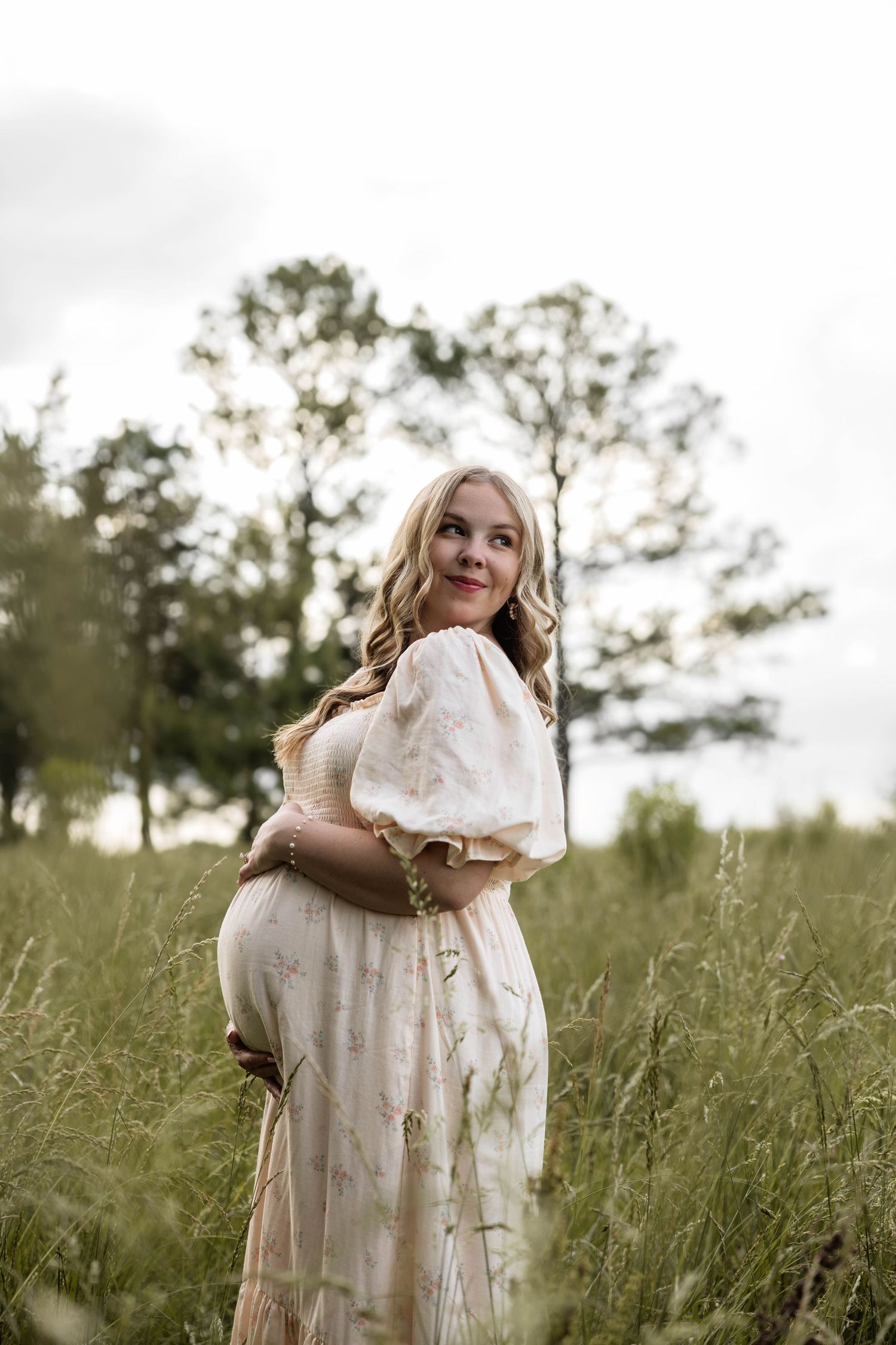 Pregnant woman in floral dress standing in tall grass field, cradling baby bump, smiling softly outdoors.