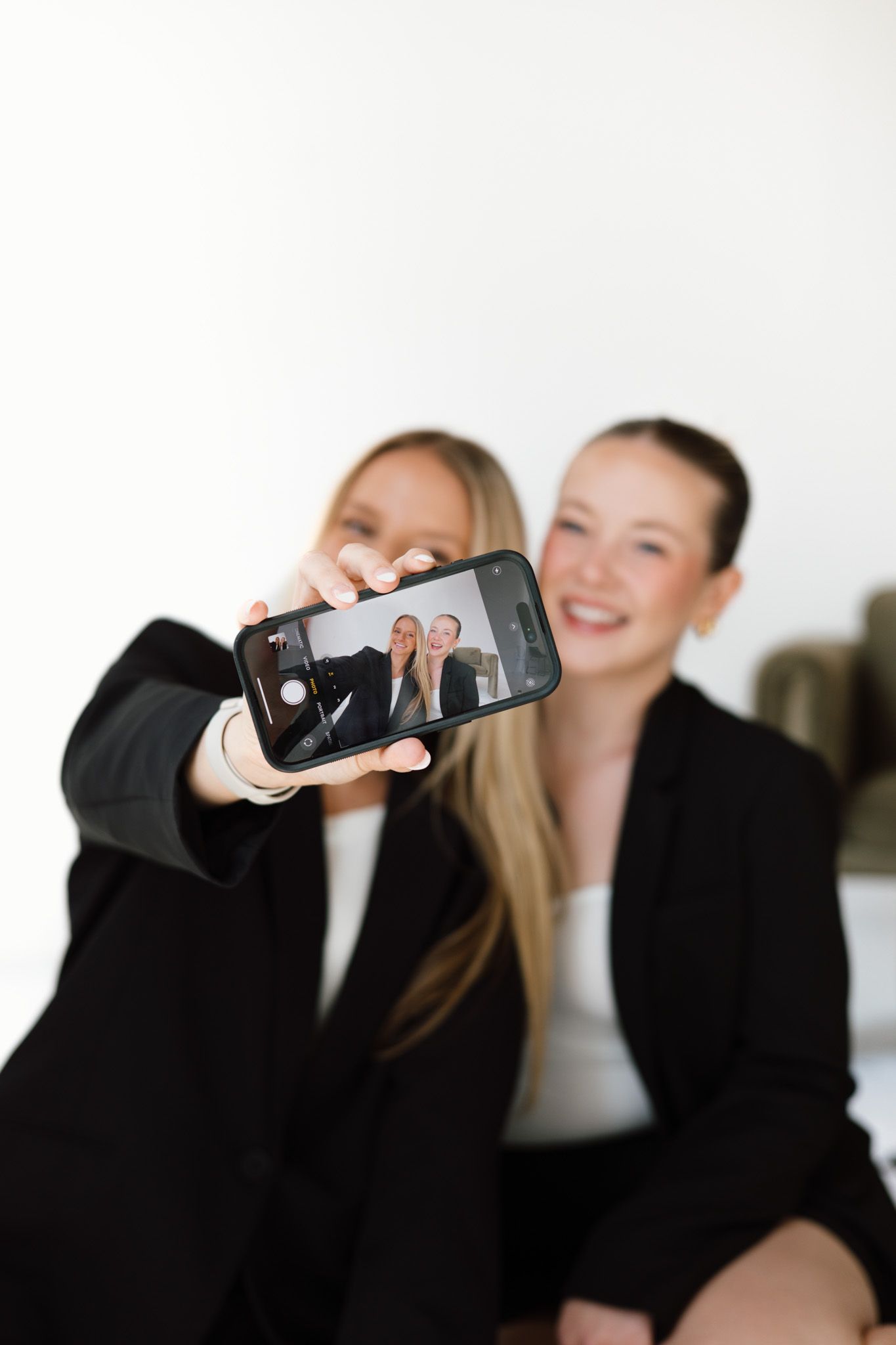 Two young women in black blazers taking a selfie together, smiling, with phone screen visible in foreground.
