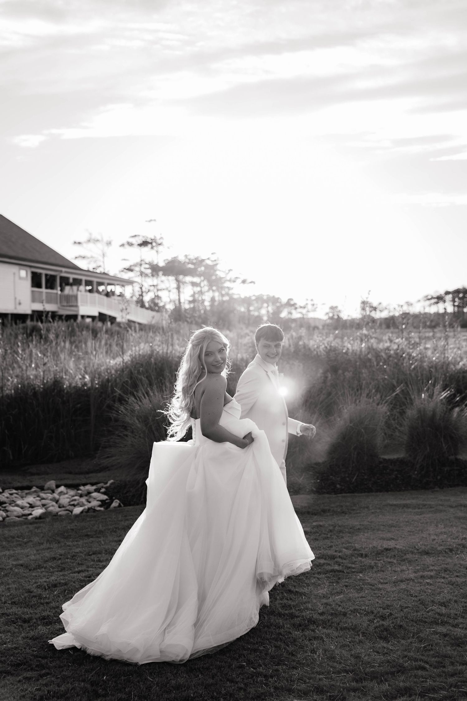 Black and white photo of a bride and groom walking outdoors at sunset near tall grass and a historic building.