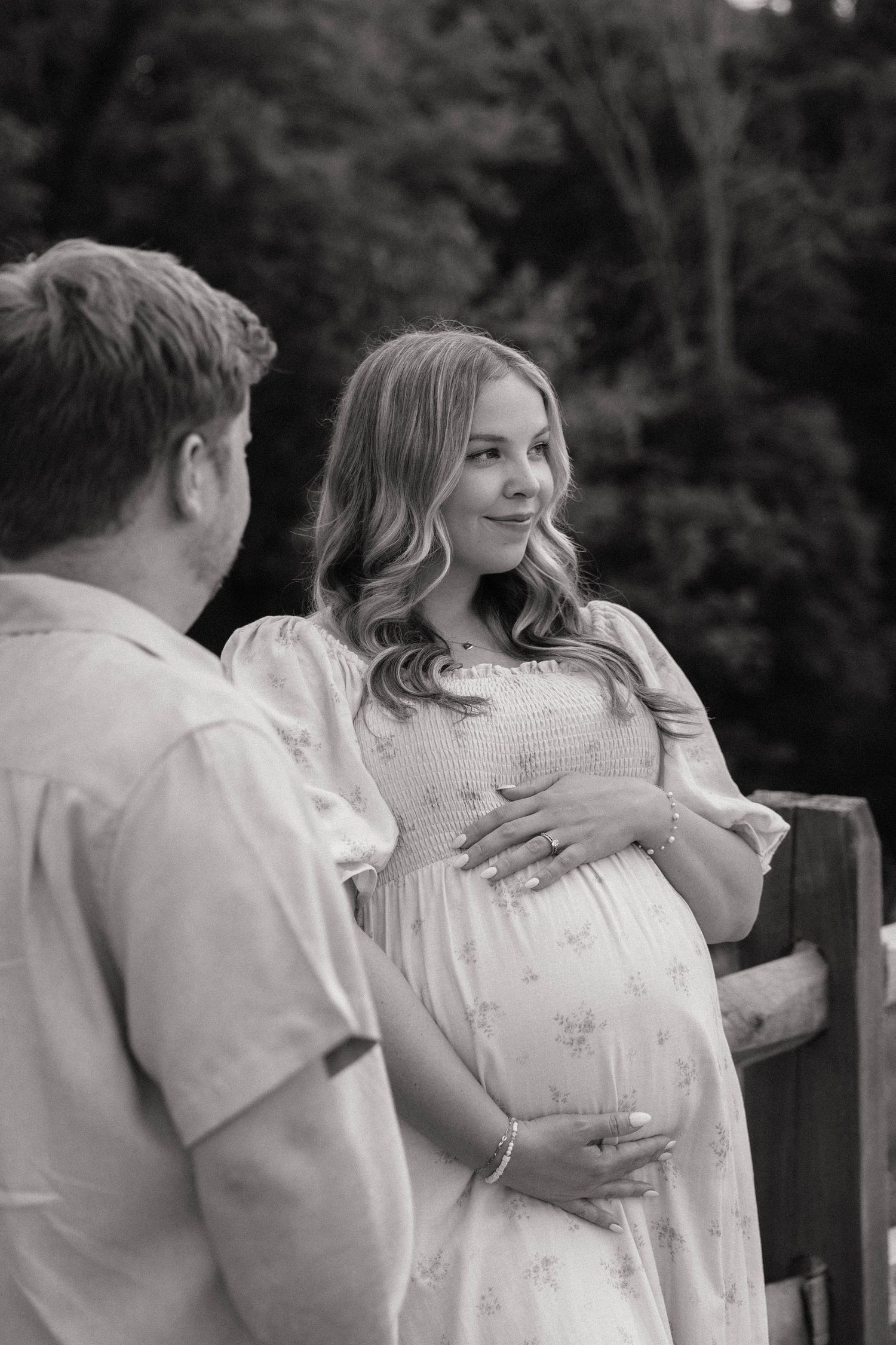 Black and white maternity photo of a pregnant woman in a floral dress smiling at her partner outdoors.