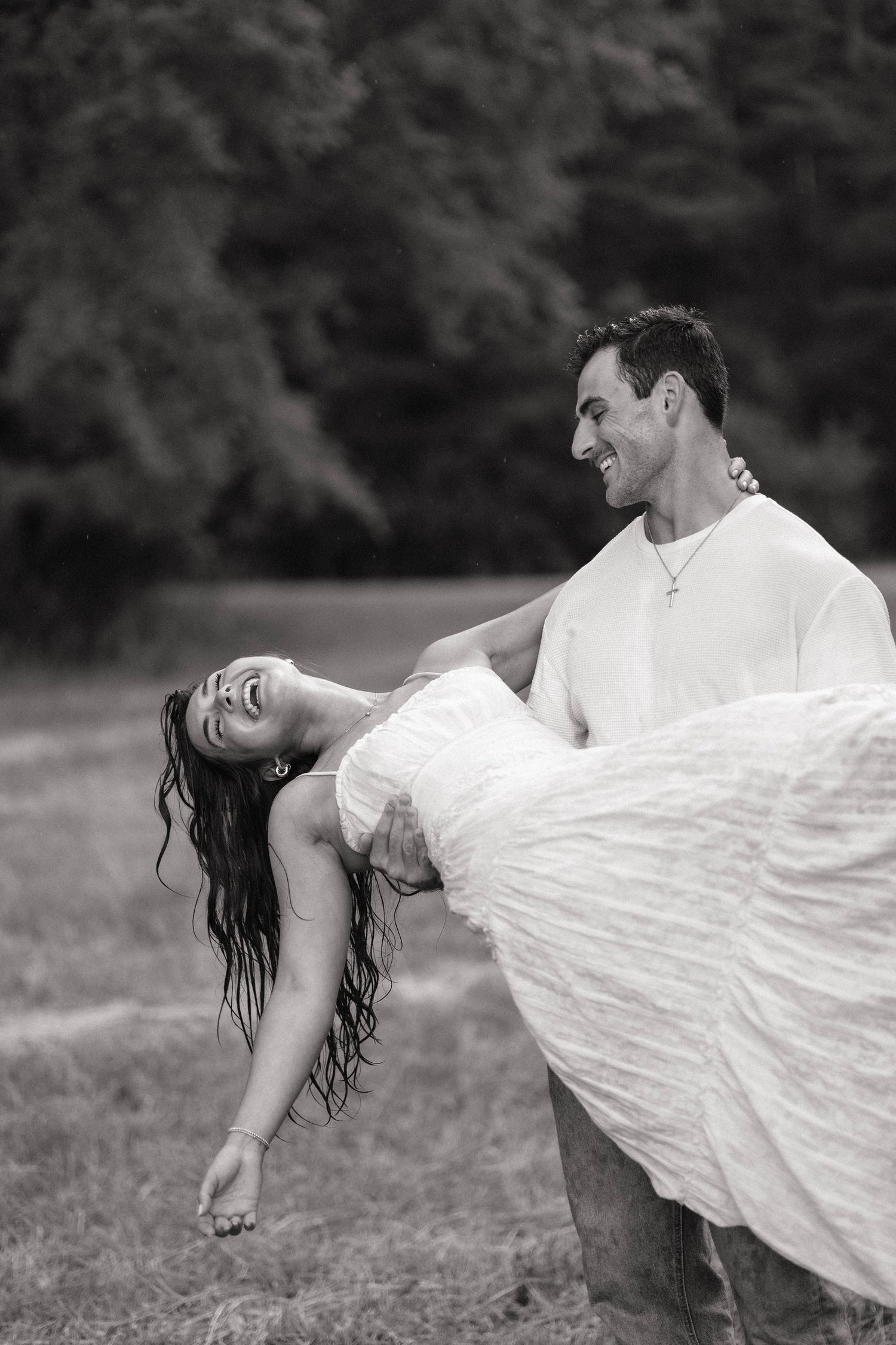 Black and white photo of a groom carrying his laughing bride outdoors, both smiling joyfully.