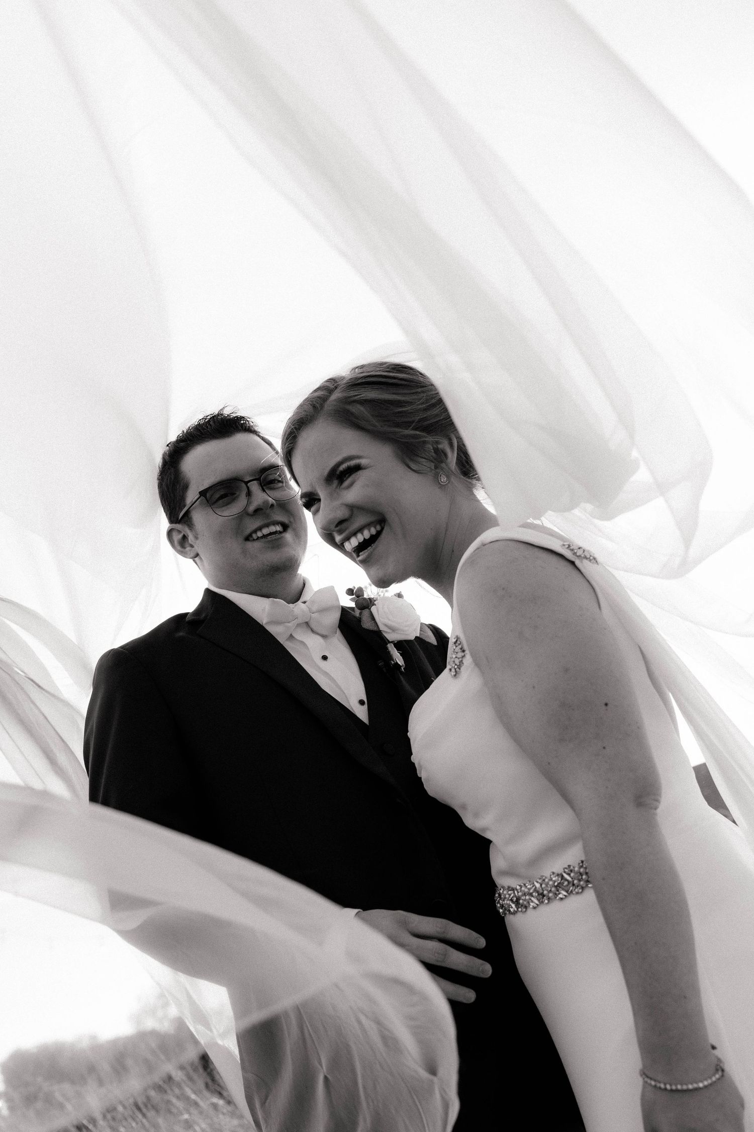 Black and white photo of a laughing bride and groom embracing under flowing white fabric on their wedding day.