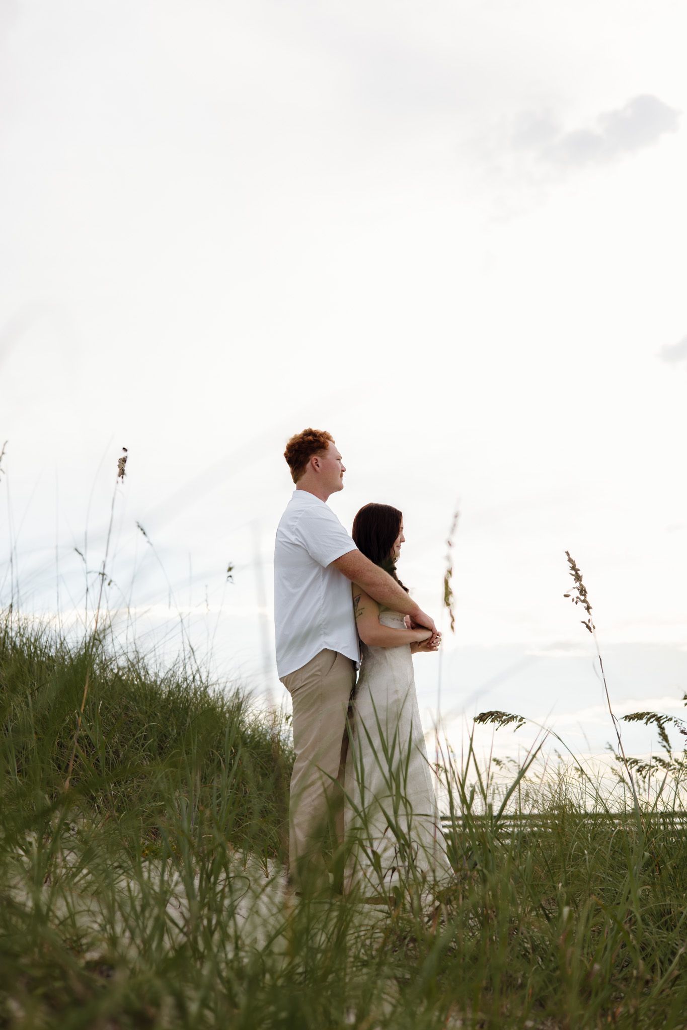 A couple embraces on a grassy dune, gazing at the horizon during a serene beach sunset.
