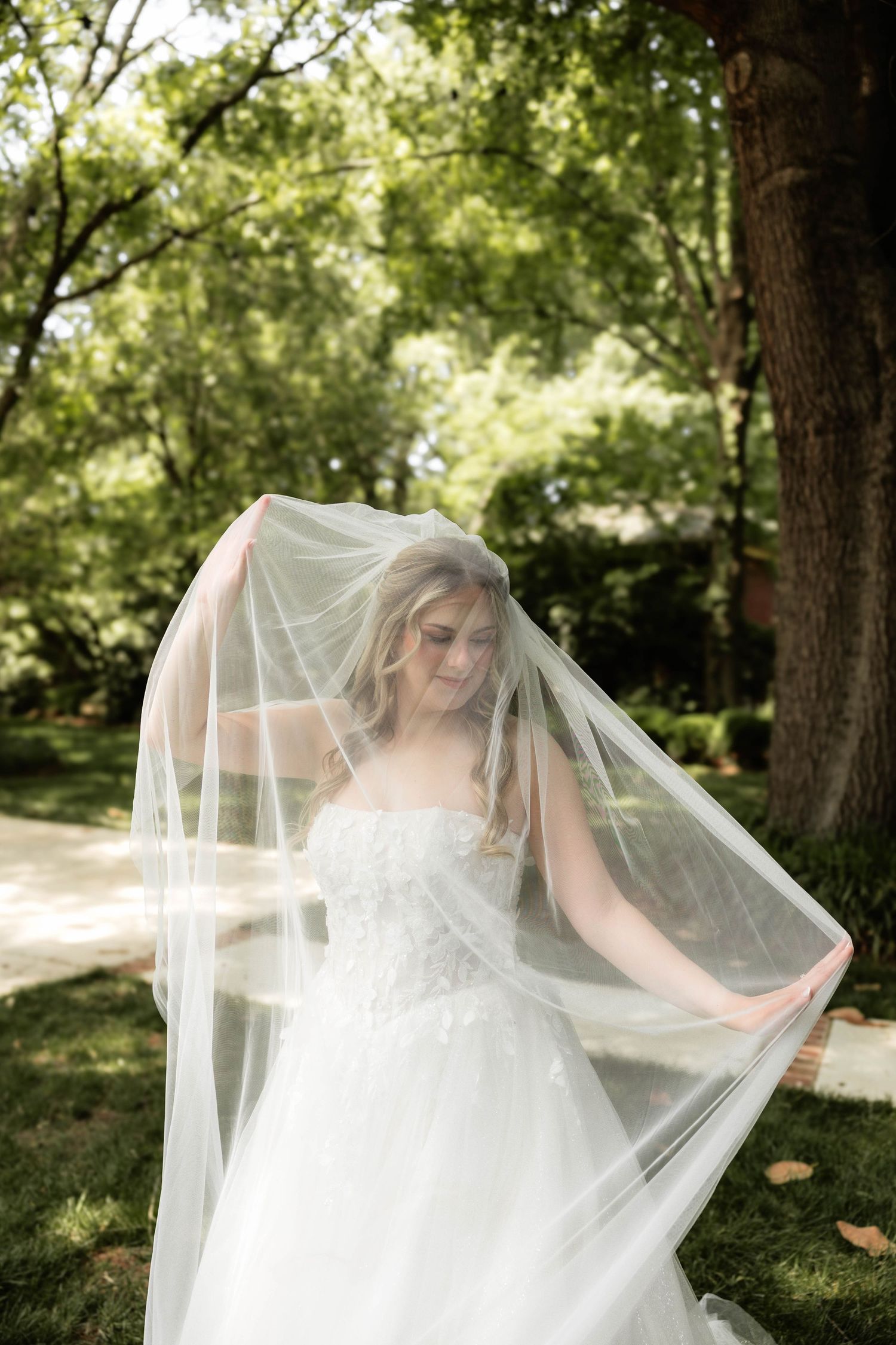Bride in white strapless gown with flowing veil lifted overhead, standing outdoors among lush green trees.