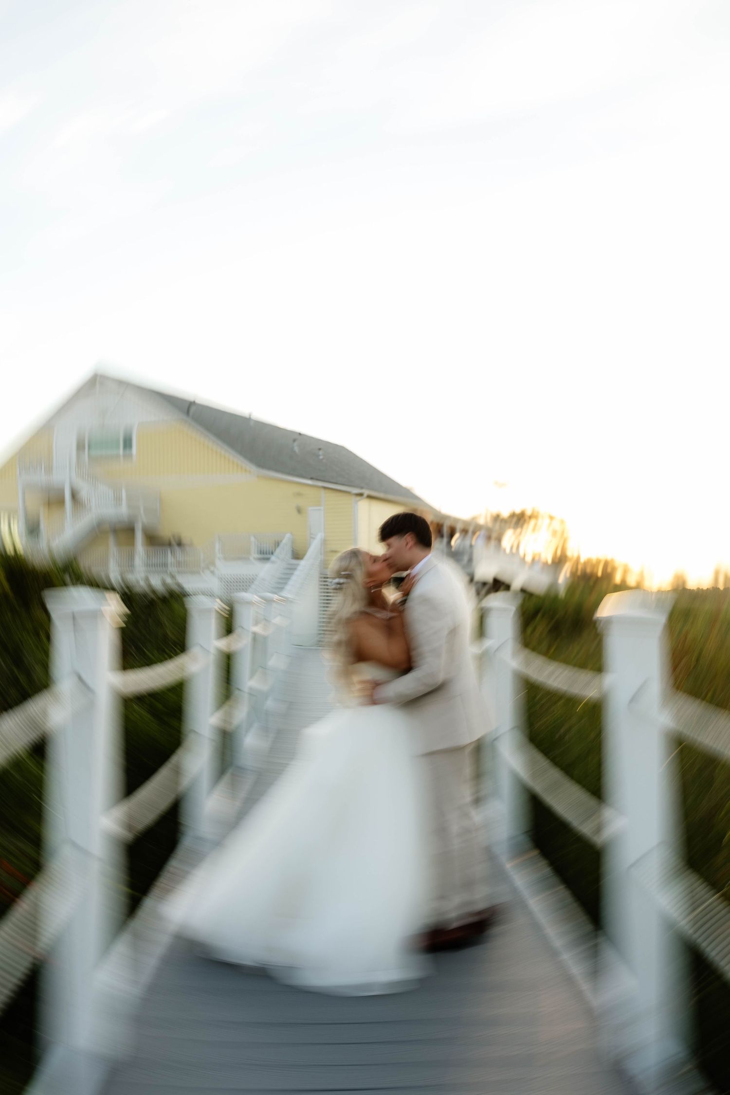 Bride and groom share a romantic kiss on a white wooden boardwalk at sunset near a yellow coastal home.