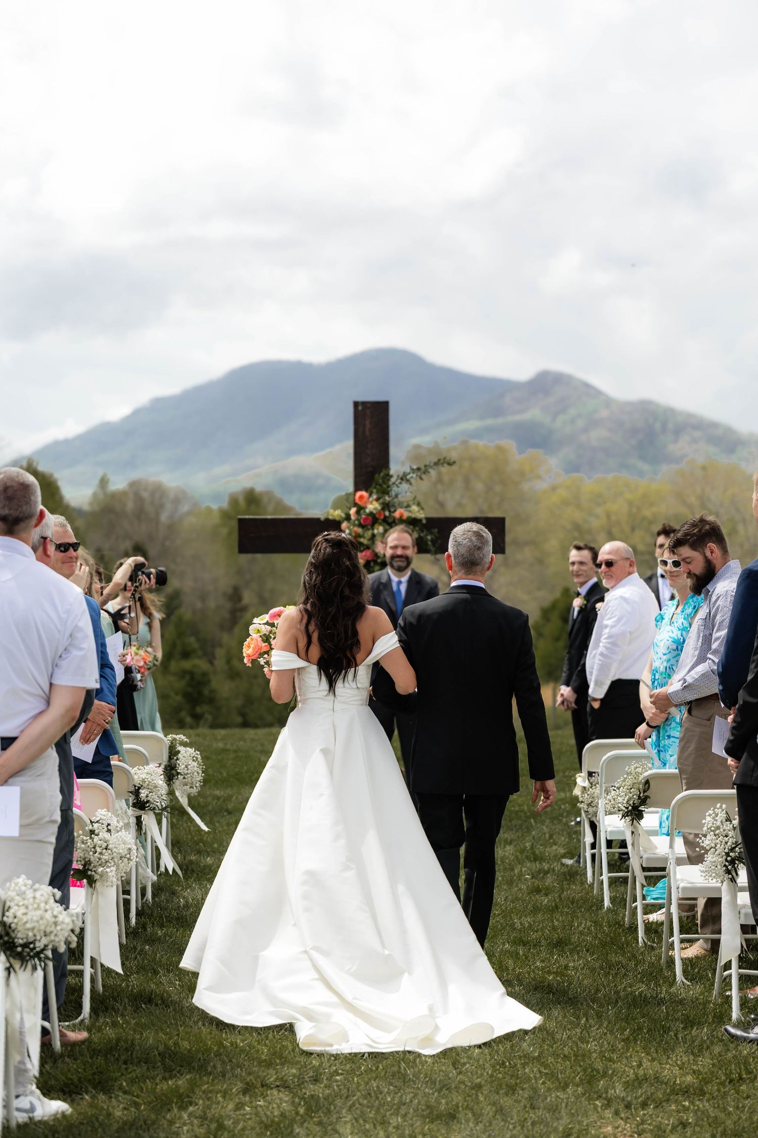 Bride walking down outdoor aisle with mountain backdrop and wooden cross at Christian wedding ceremony.