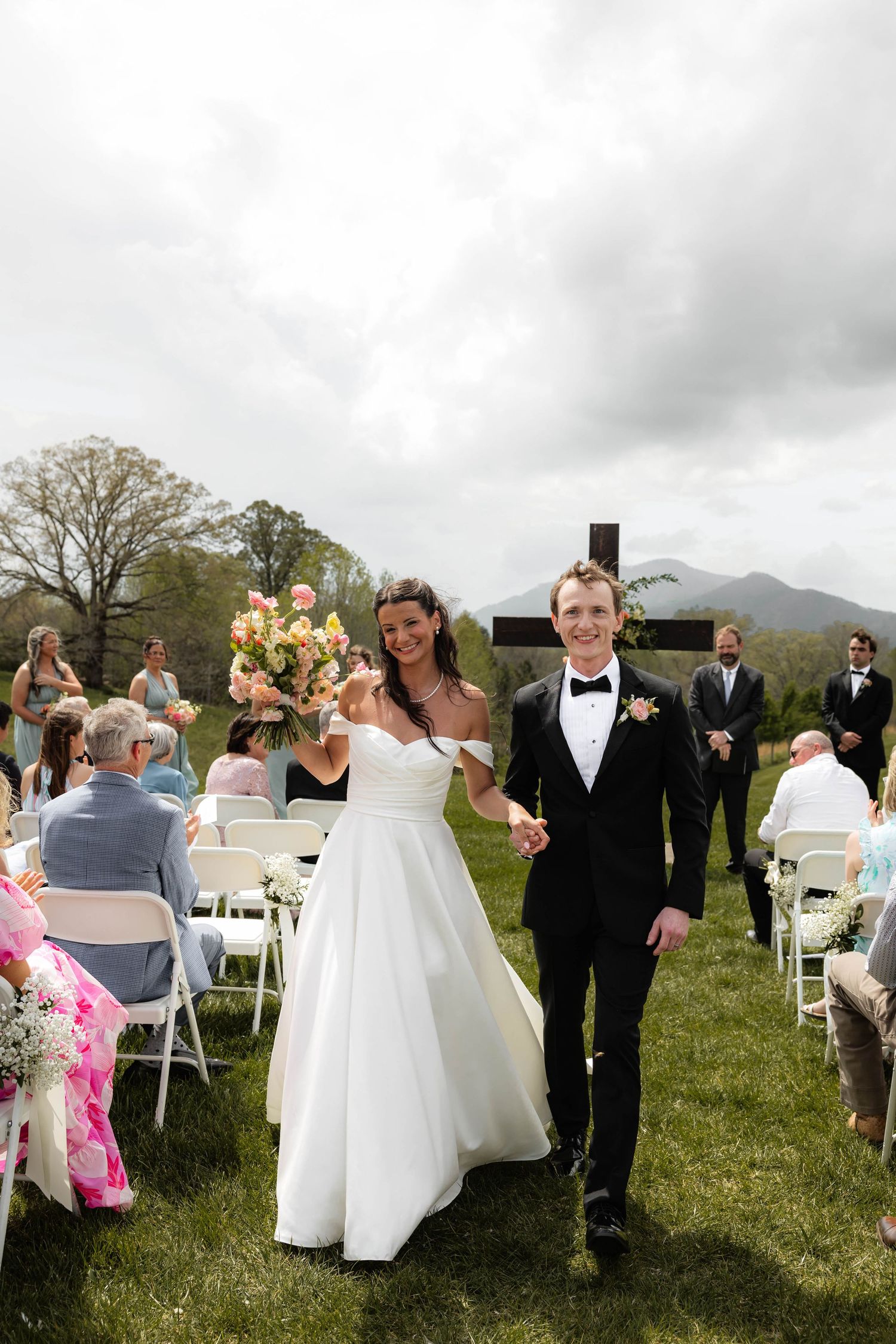 Bride and groom walk down outdoor aisle after wedding ceremony, mountains in background, bride holding pink bouquet.