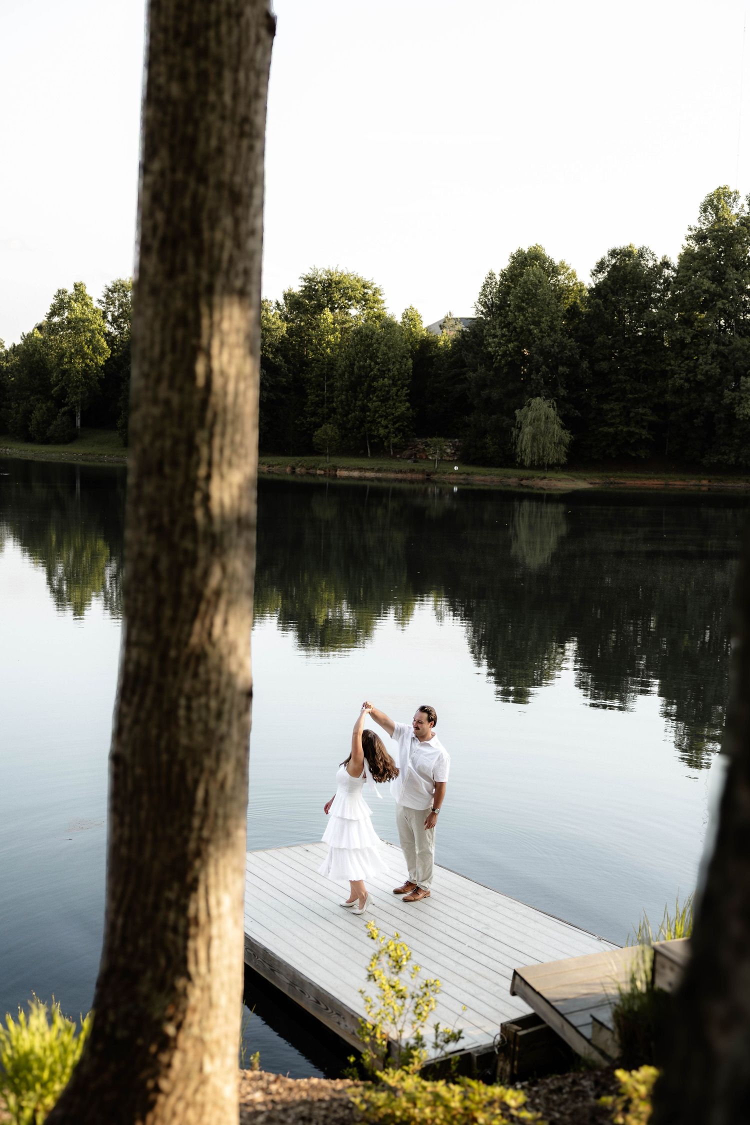 Couple in white dancing on a wooden dock by a calm reflective lake surrounded by lush green trees.