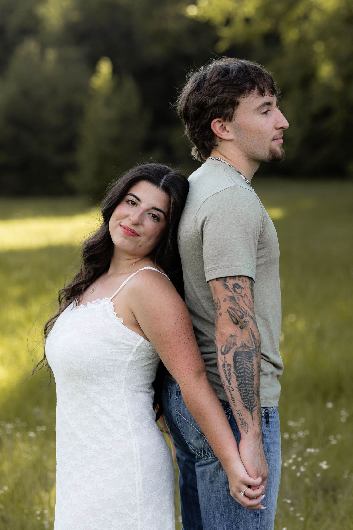 Couple standing back-to-back in a green field, woman in white dress smiling, man with tattoo sleeve in jeans.