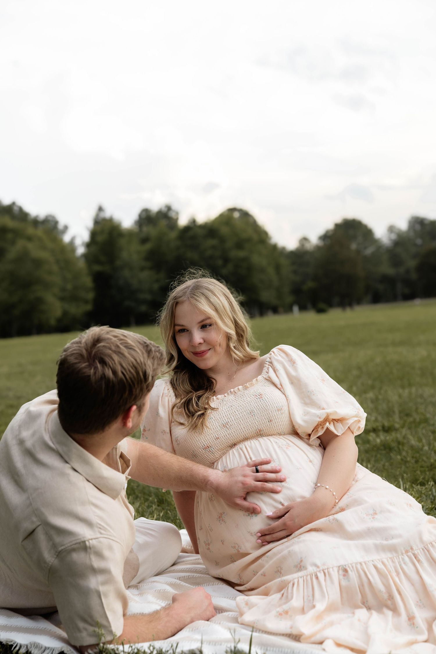 Couple sitting in a green field, man tenderly touching pregnant woman's belly during outdoor maternity photo session.