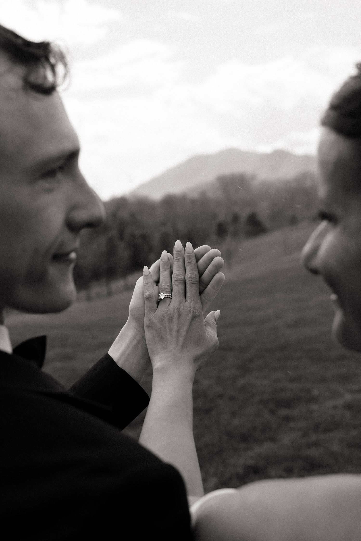 Black and white photo of a smiling couple holding hands, showcasing an engagement ring, with mountains in the background.