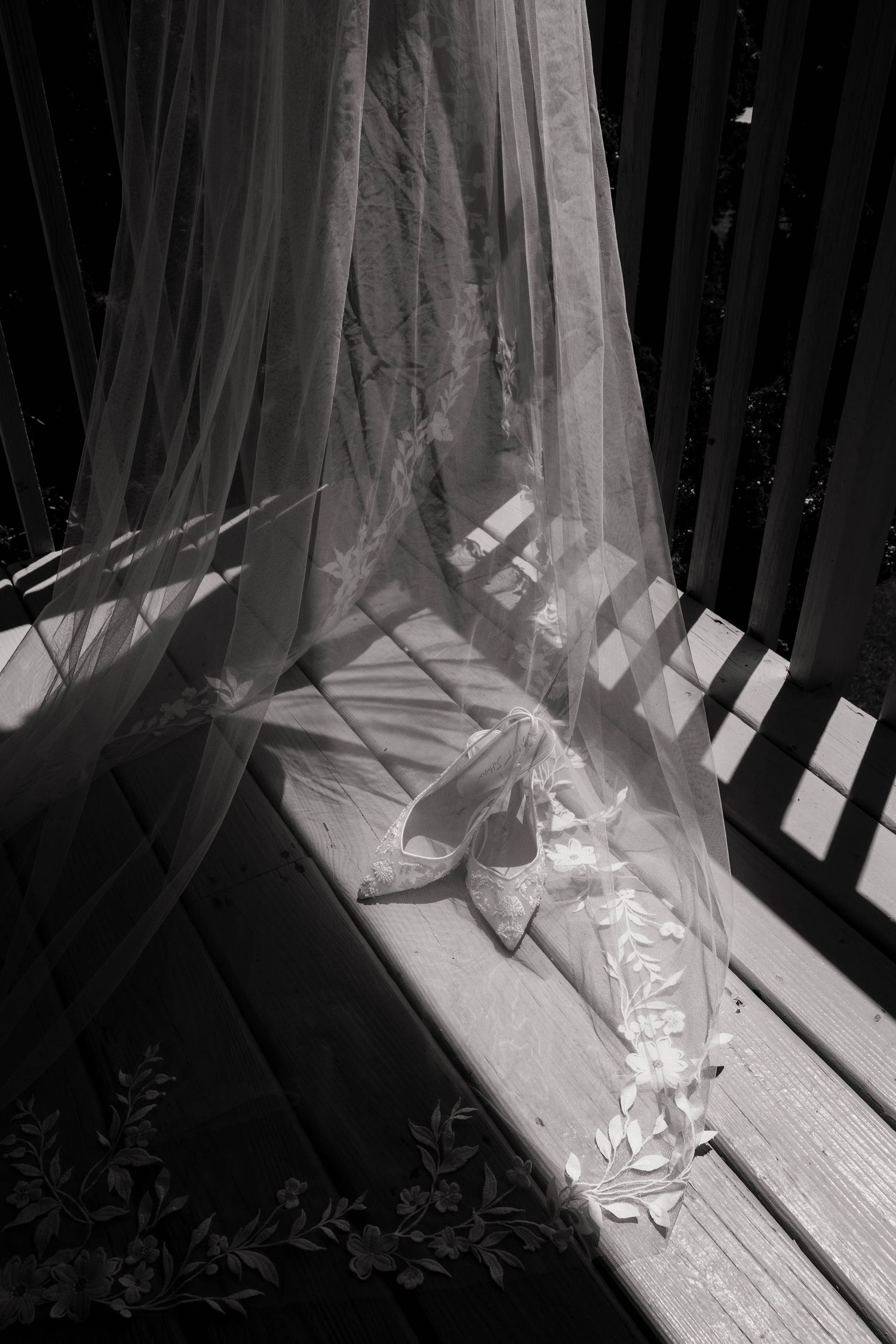 Black and white photo of bridal veil and wedding shoes on wooden bench with sunlight shadows.