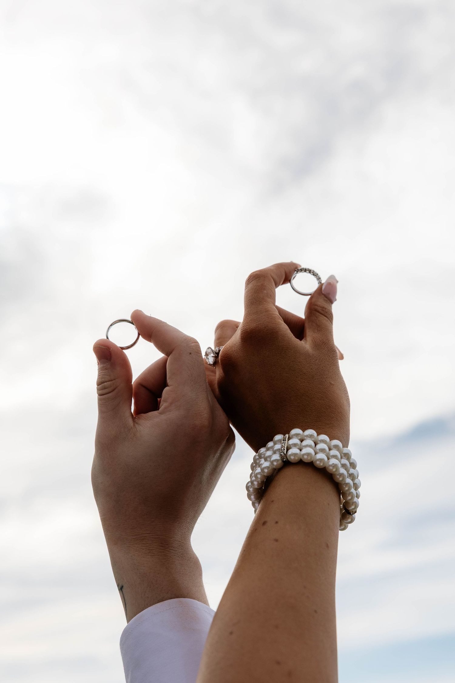 Two hands holding wedding rings up against a cloudy sky, one wrist adorned with a pearl bracelet.