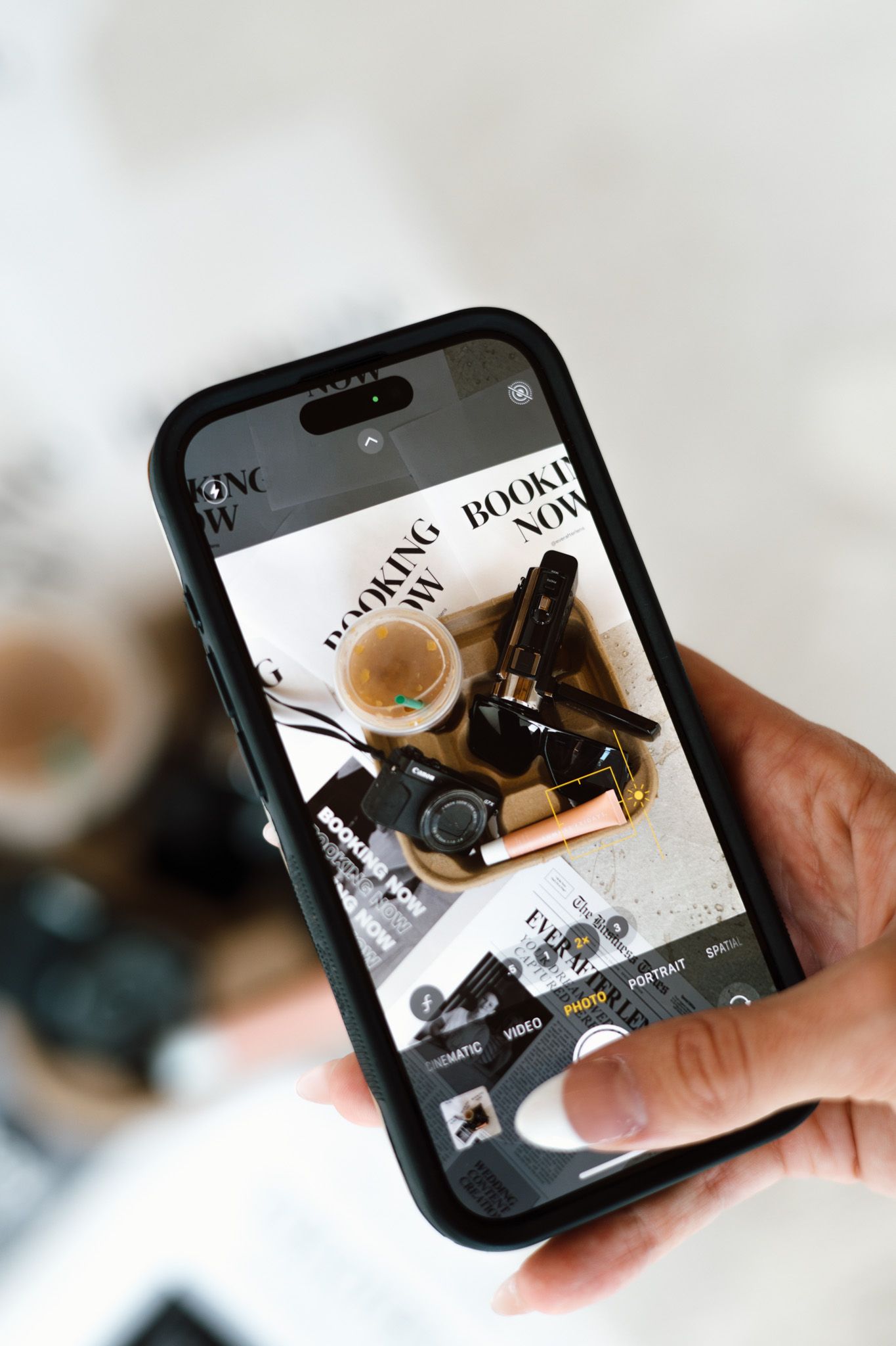 Hand holding smartphone photographing flat lay of makeup products, camera, and coffee on white surface.