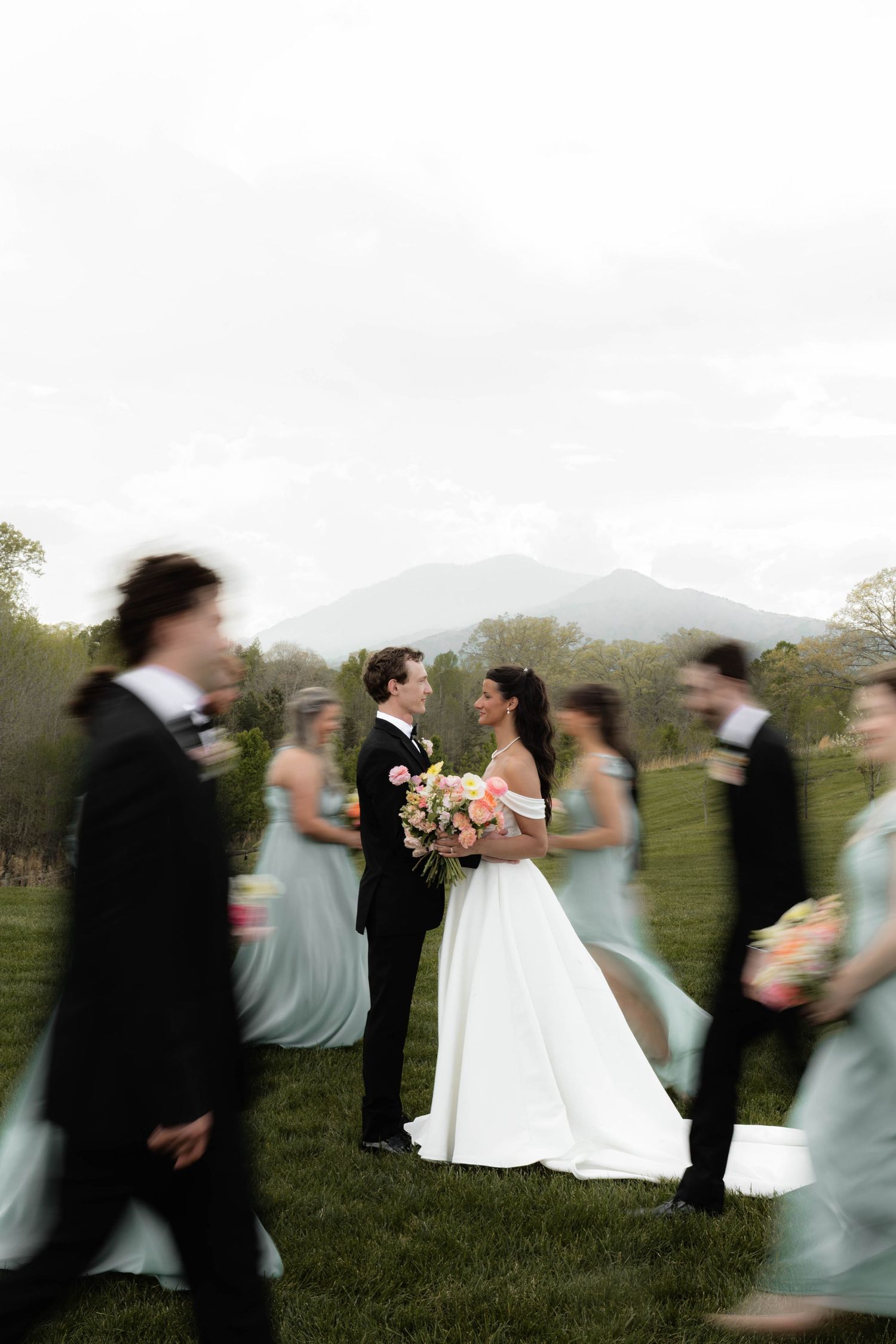 Bride and groom share a romantic moment as blurred wedding party walks past, with misty mountains in the background.