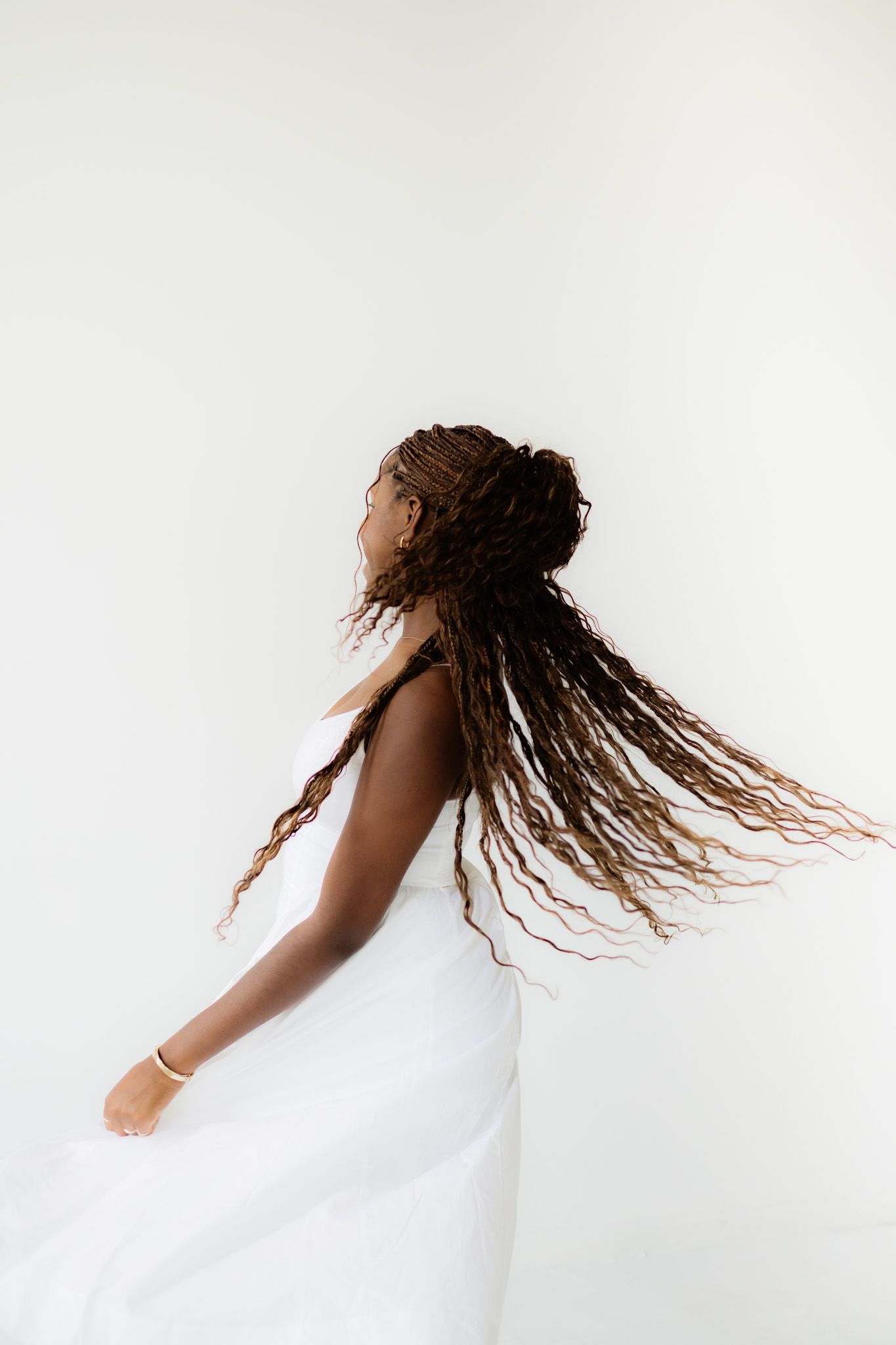 A Black woman with long braided locs tosses her hair while wearing a white dress against a white background.