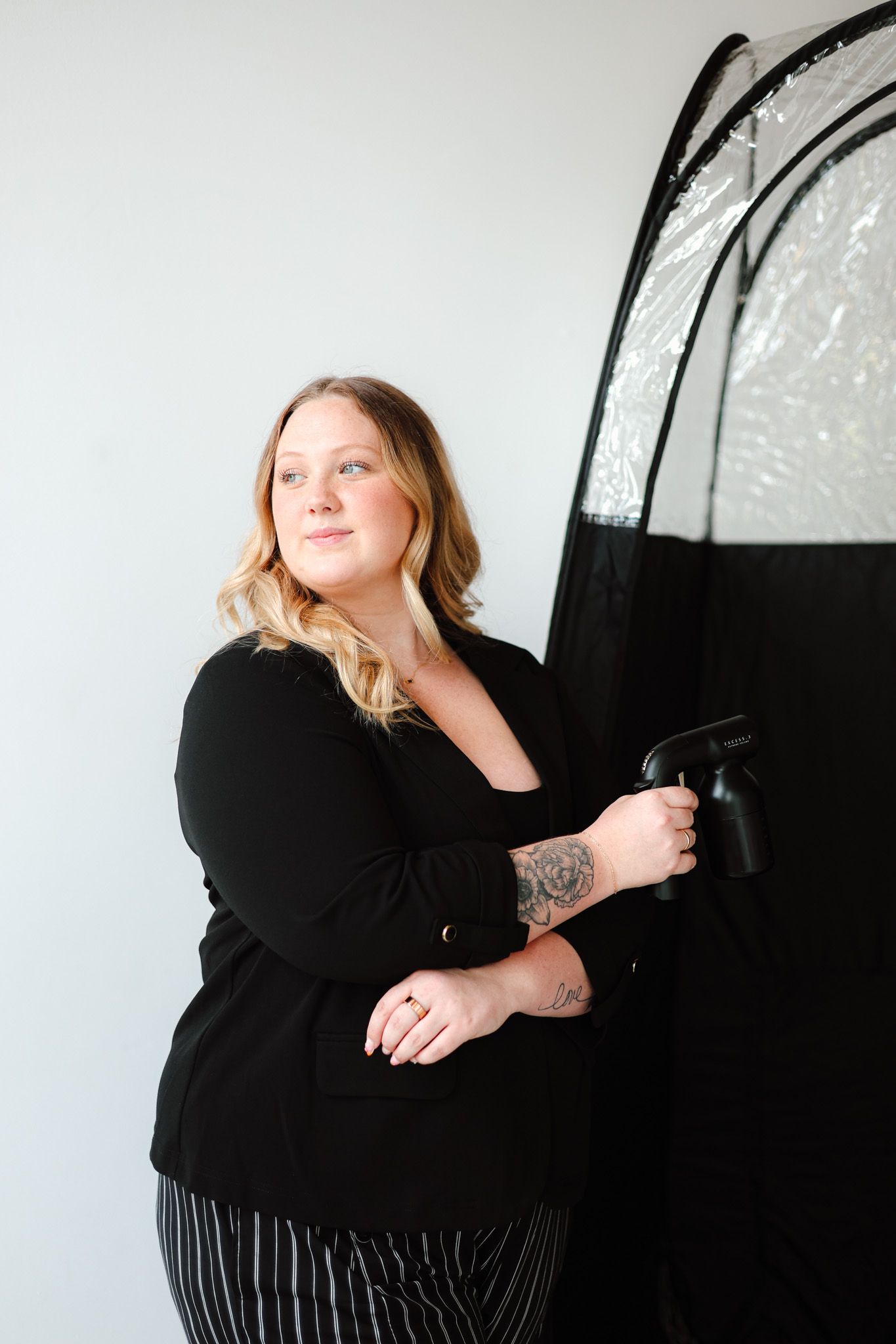 Woman in black blazer and striped pants holding photography equipment next to a reflective softbox in a studio.