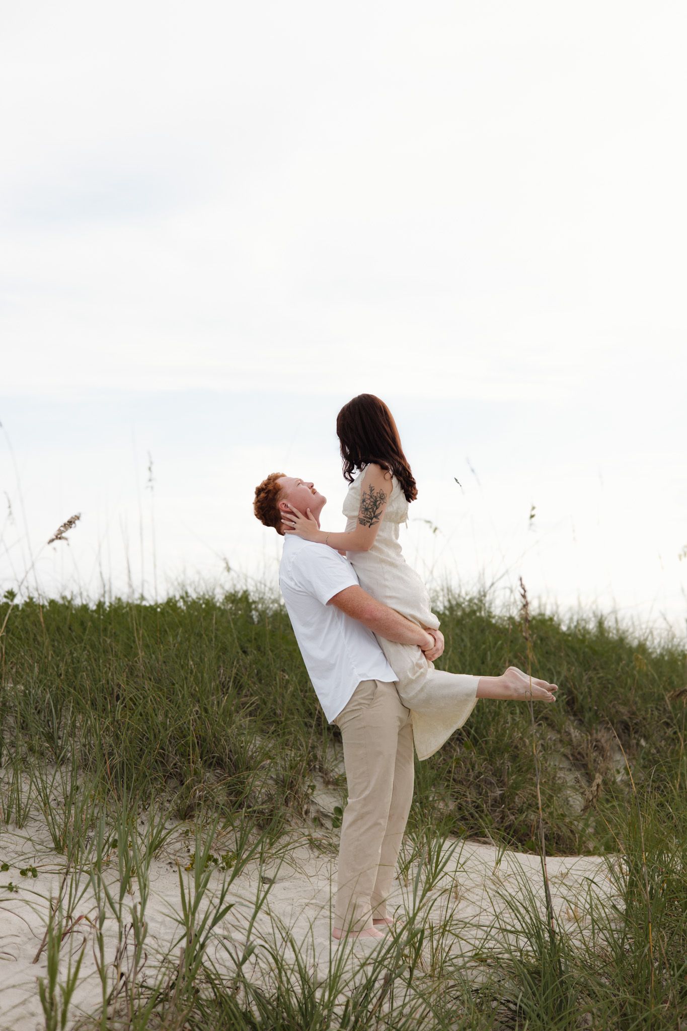 A man lifts a woman on sandy beach dunes with sea oats, both dressed in white and beige, sharing a romantic moment.