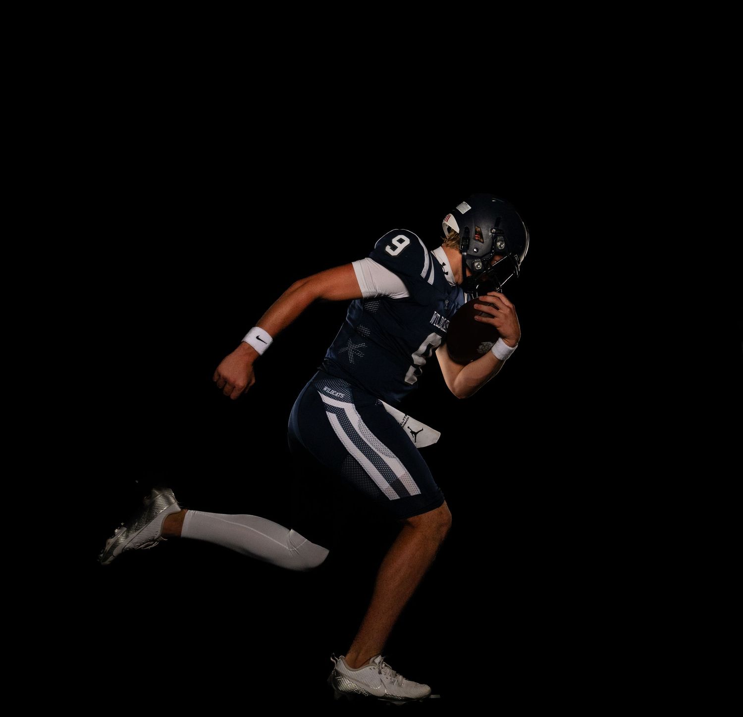 Football player in dark uniform runs dynamically against black backdrop.