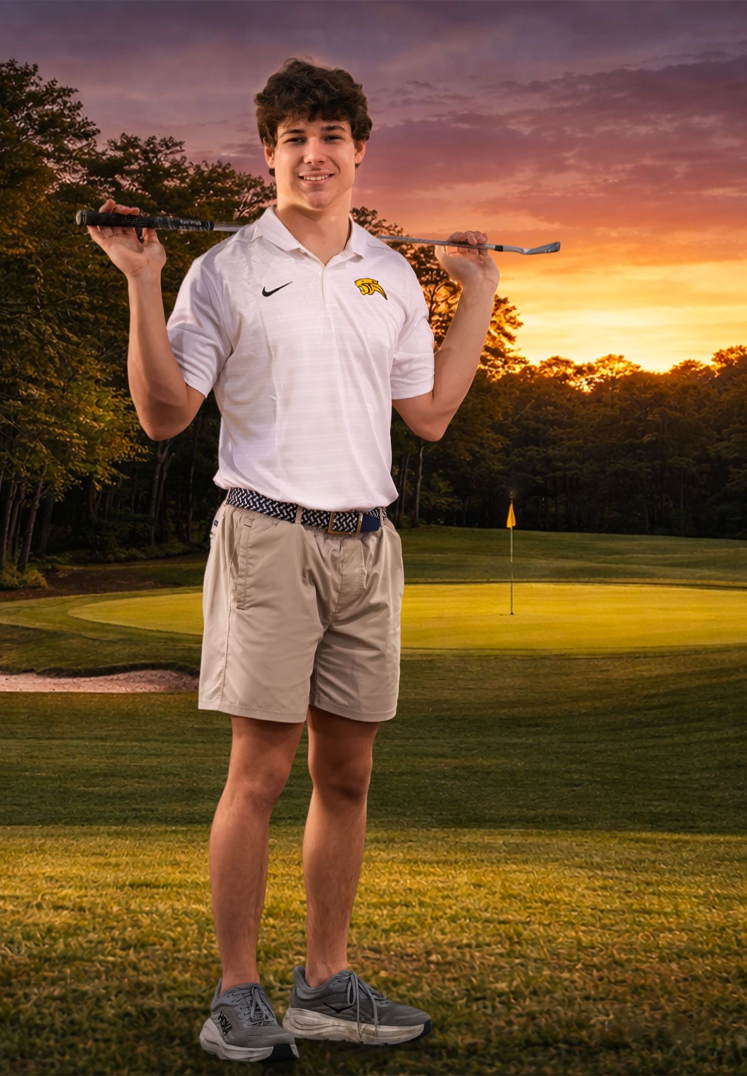 Student athlete in Nike polo holds golf clubs on course during dramatic sunset.