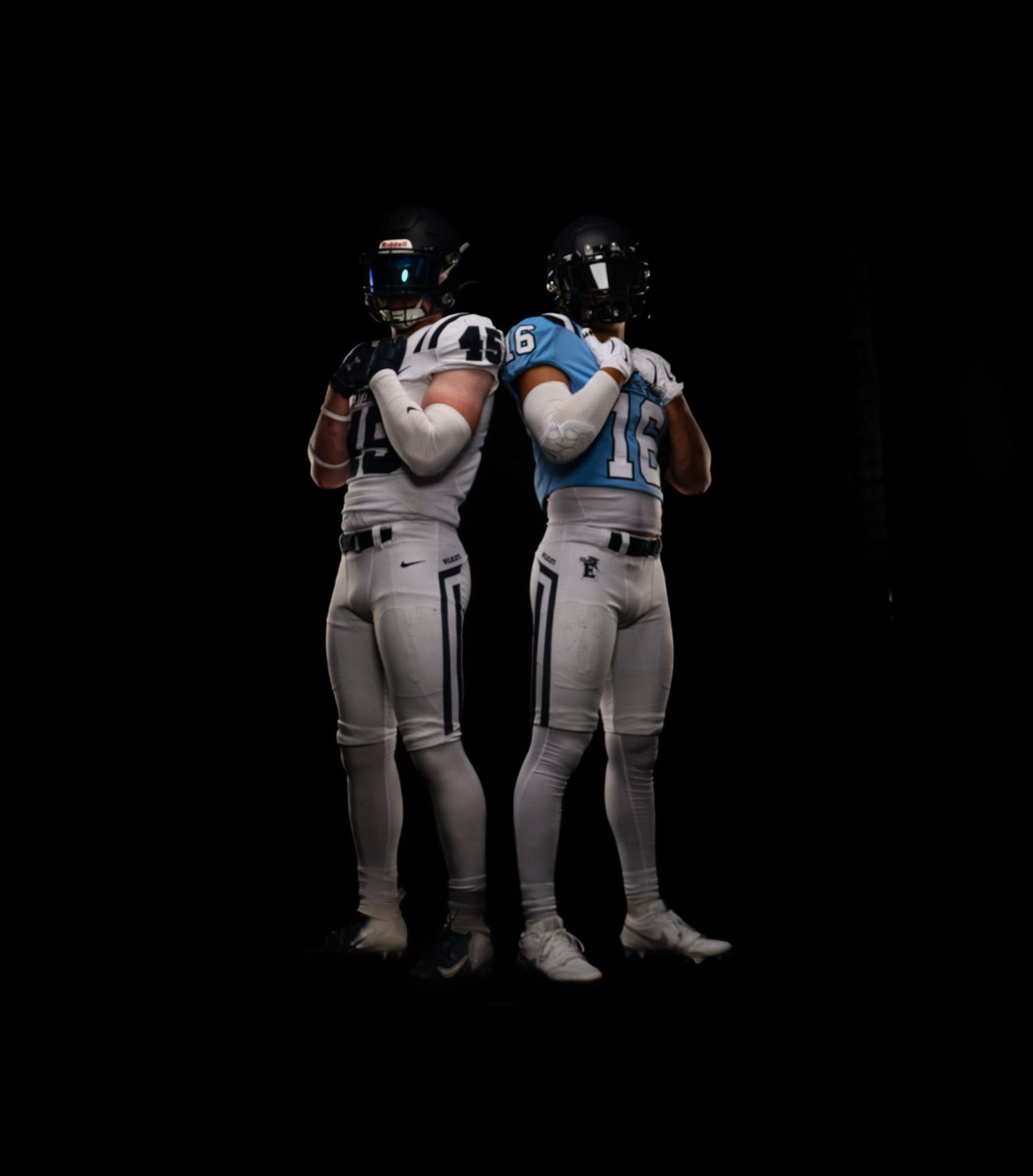 Two football players in white uniforms and helmets pose back-to-back against a dark background.