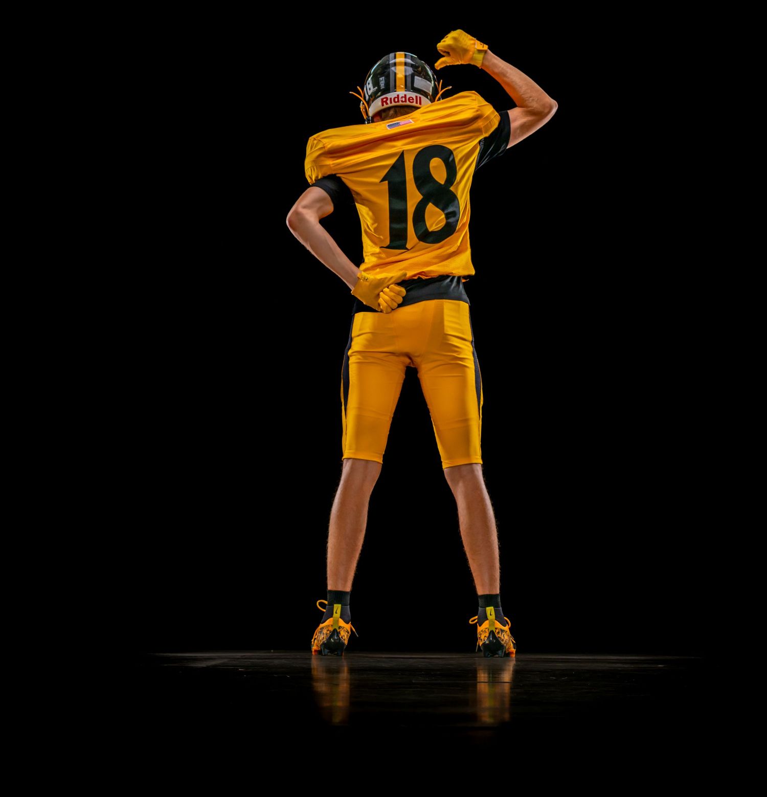 Football player number 18 in gold uniform flexing from behind against black background.
