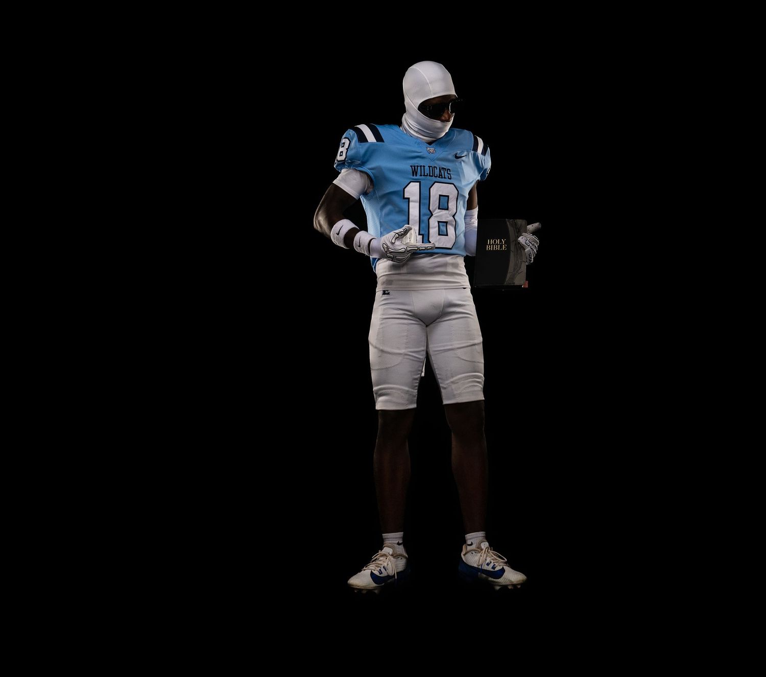 Football player in blue #18 jersey with white face mask holds item against dark background.