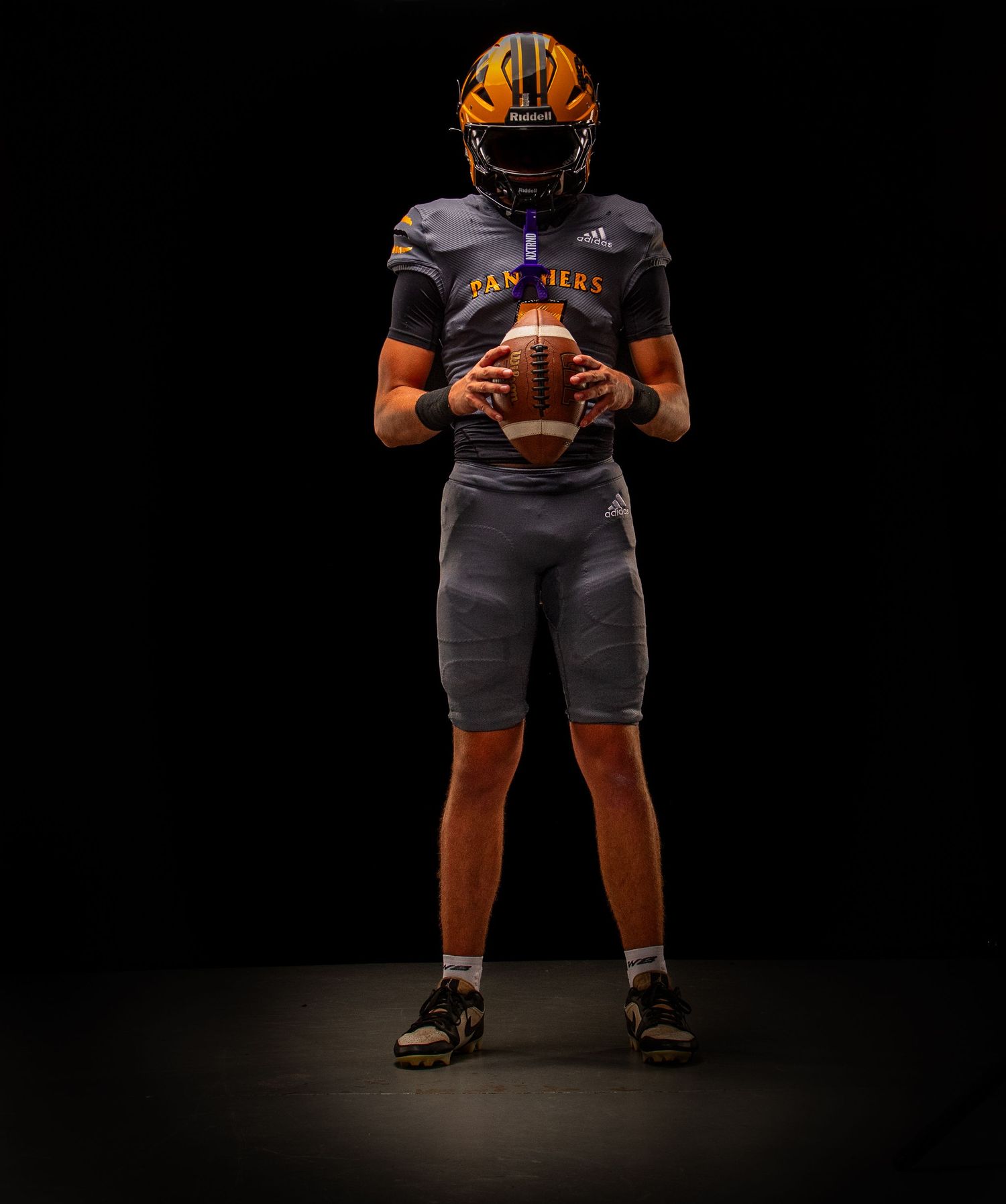 Football player in gray and gold uniform holding a football, wearing an orange helmet, posed against a dark background.