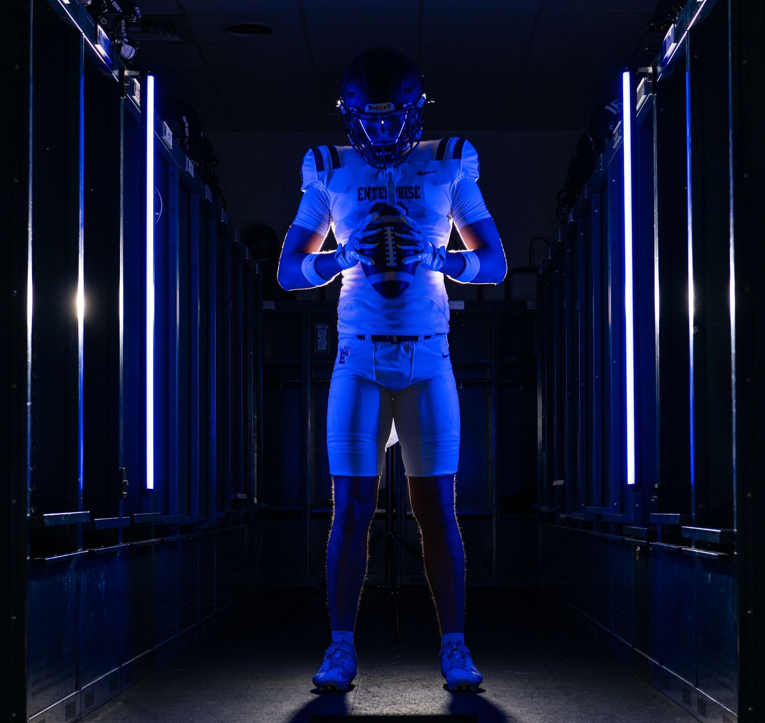 Football player in blue uniform poses dramatically in a dark locker room hallway with glowing blue LED lights.