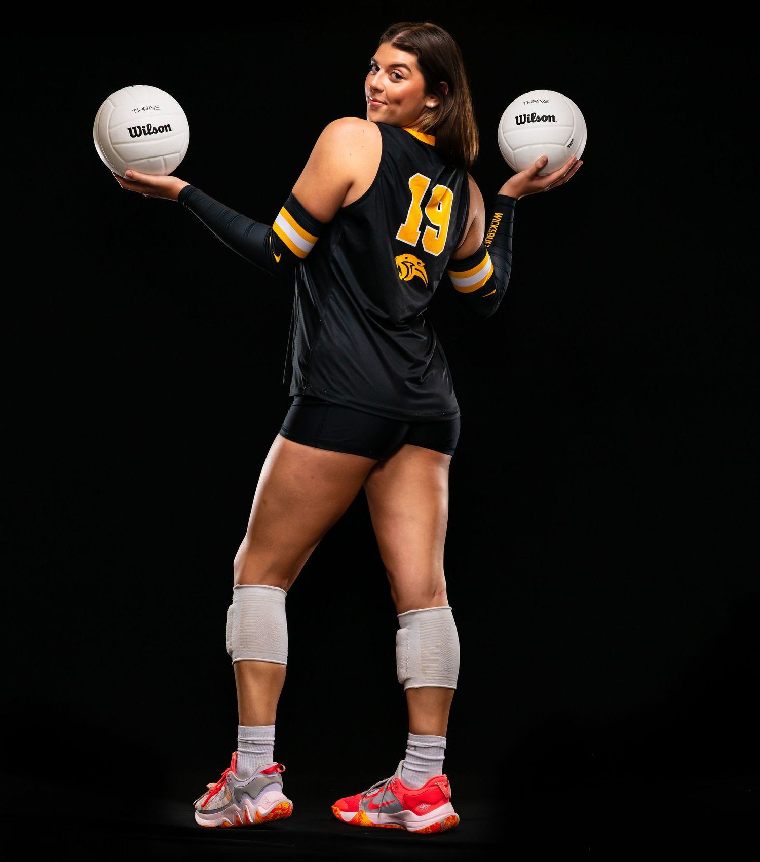 Volleyball player in black and gold jersey #19 holds two Wilson volleyballs, wearing knee pads and colorful sneakers.