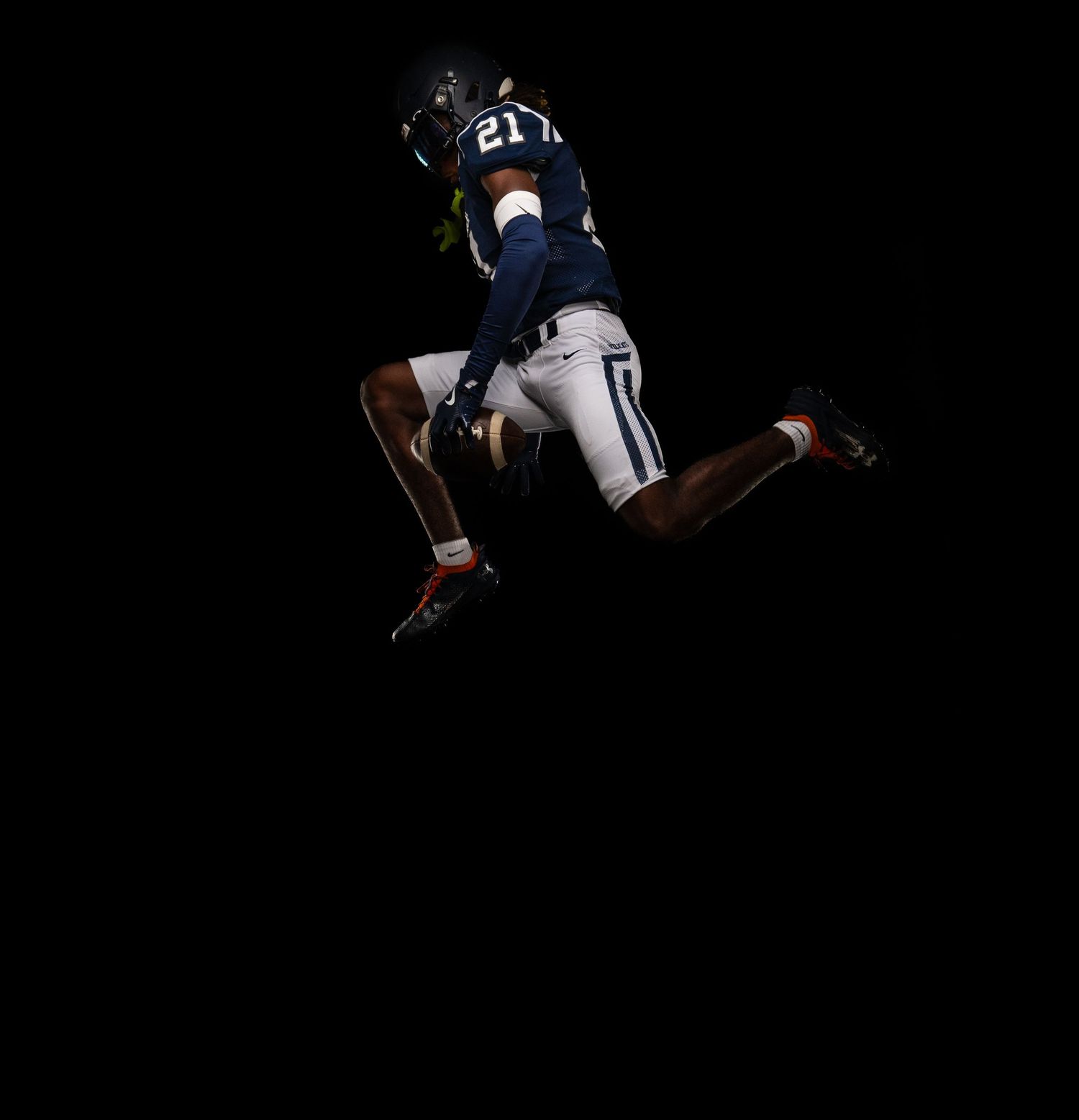 Football player in navy jersey leaps high against dark background.