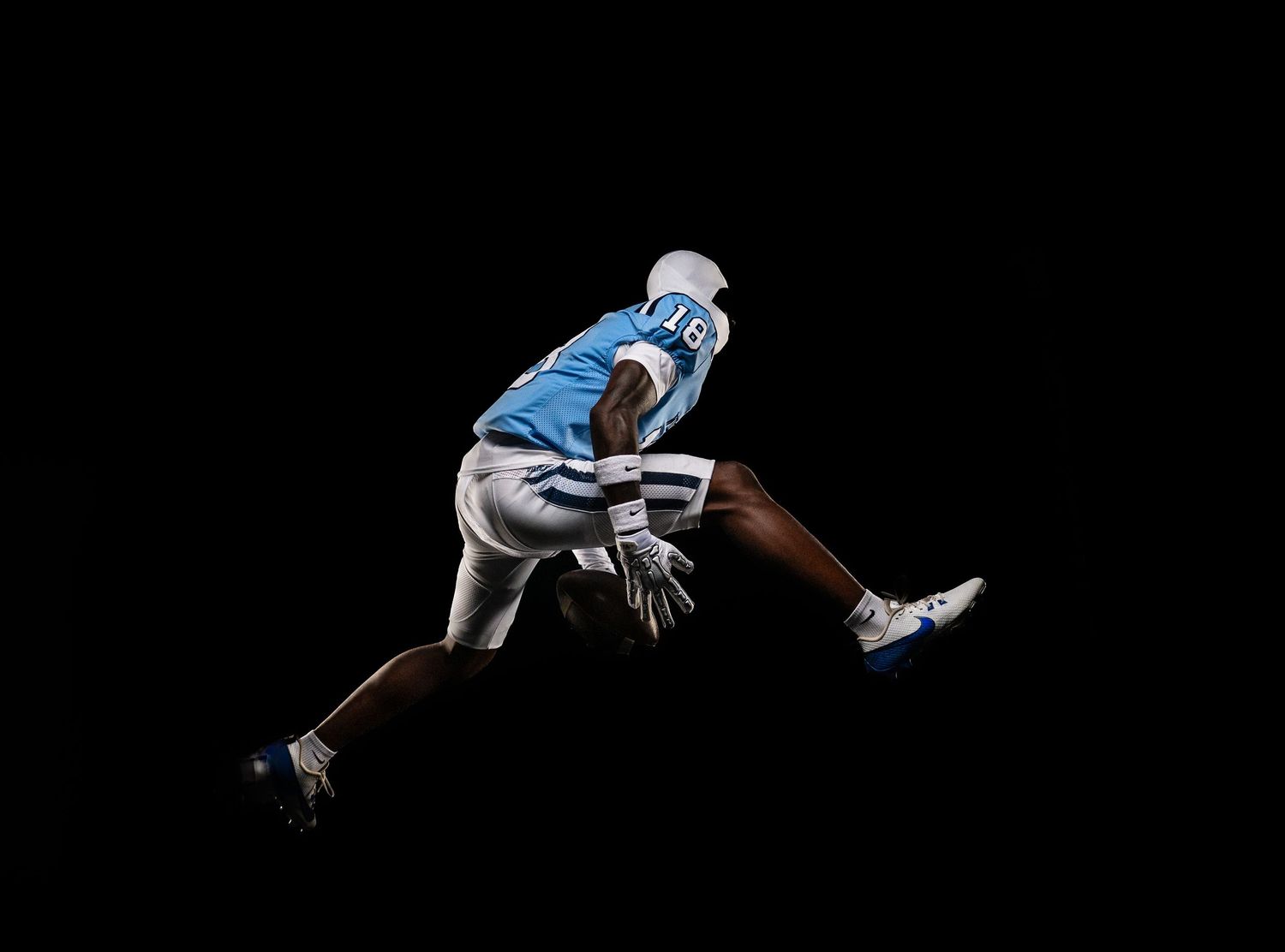 Athletic football player in light blue jersey performs dynamic stretching pose against black background.