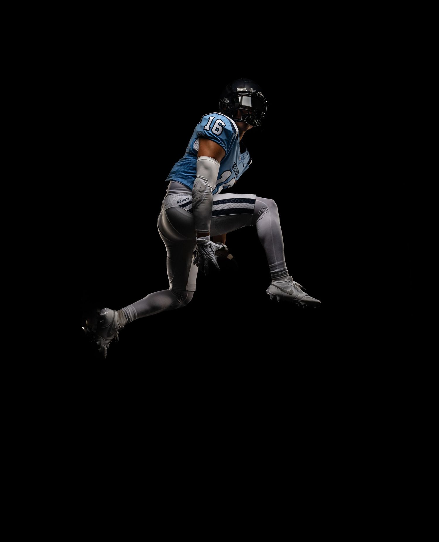 Football player number 16 in blue jersey leaps mid-air against black background.