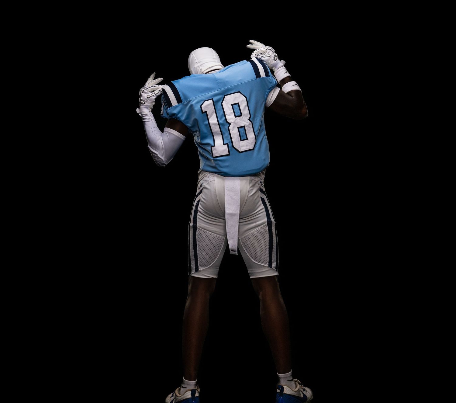 Football player wearing number 18 blue jersey and white pants, standing with back turned on dark background.