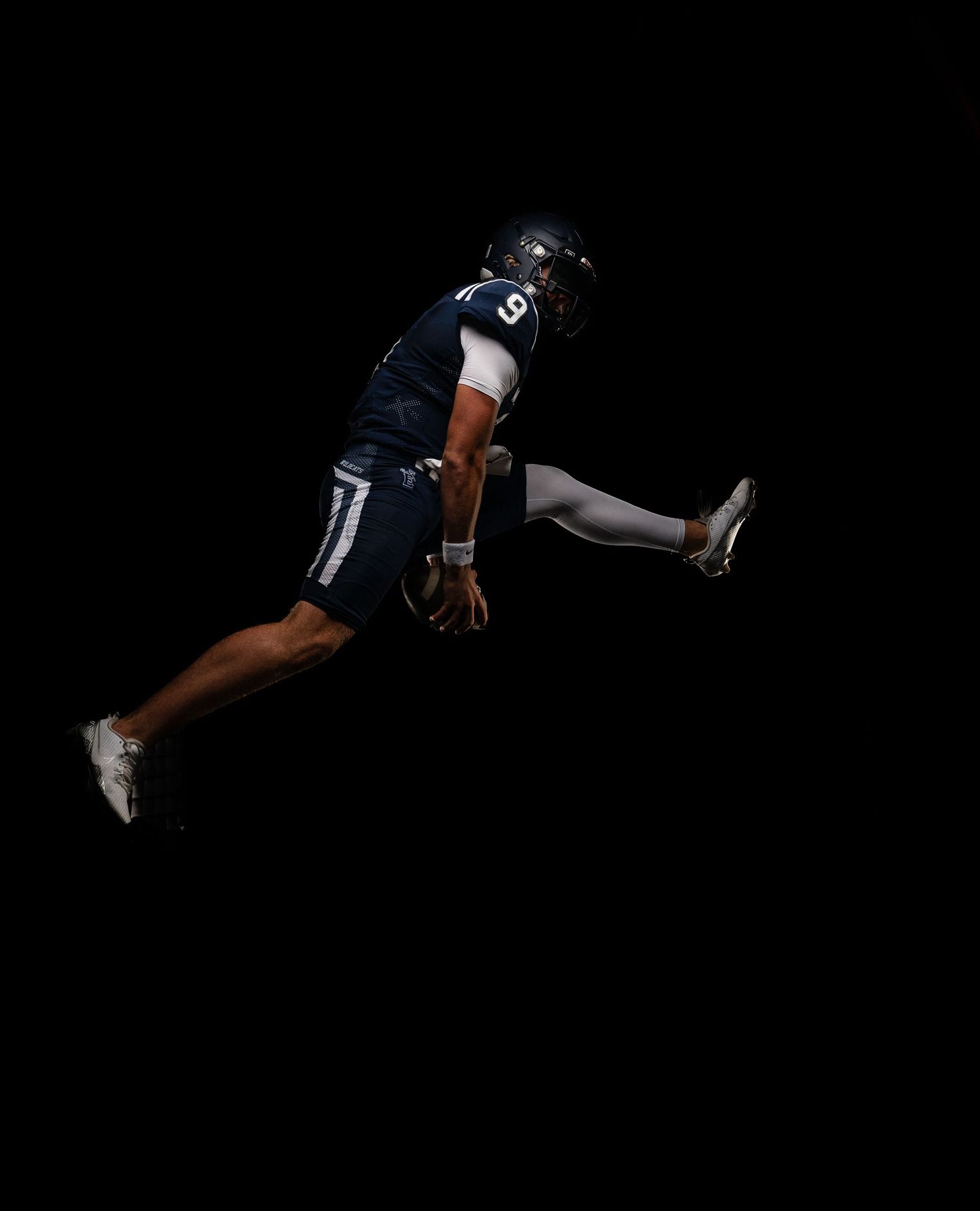 Football player in dark uniform leaping dynamically against black background, showcasing athleticism and speed.