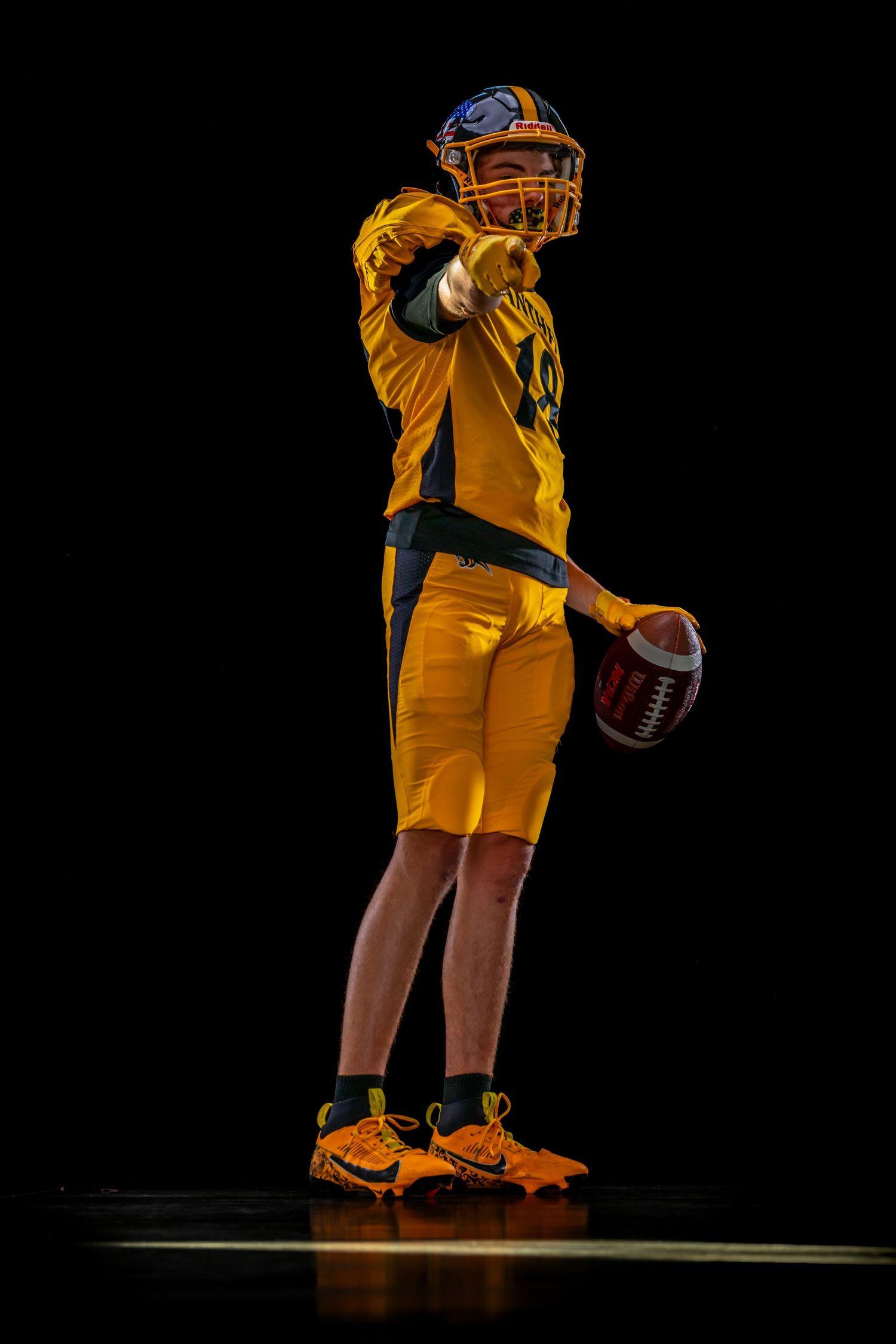 Football player #18 in yellow uniform holds ball and points forward against black background.