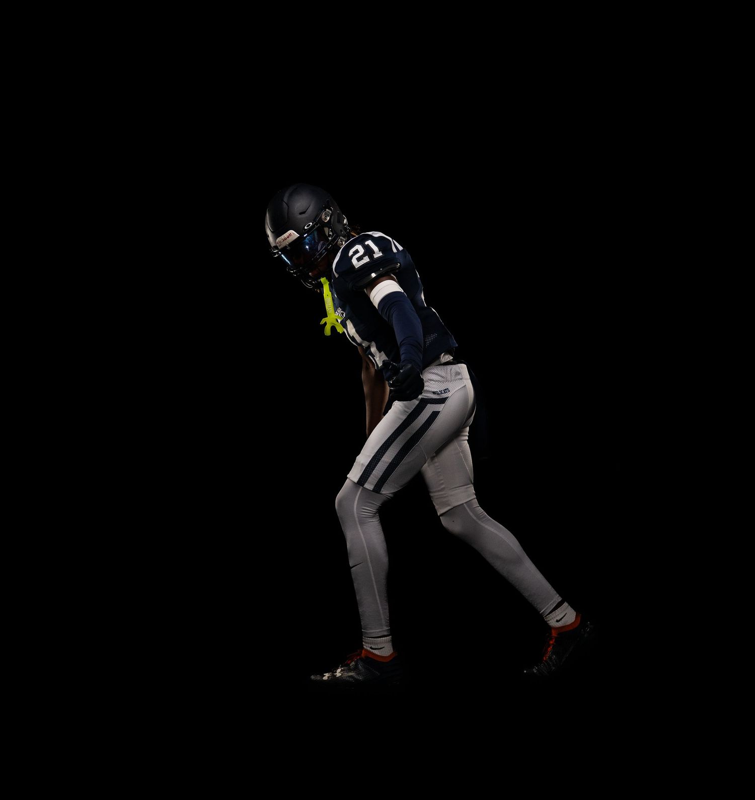Football player number 21 in navy uniform crouches in athletic stance on dark background.