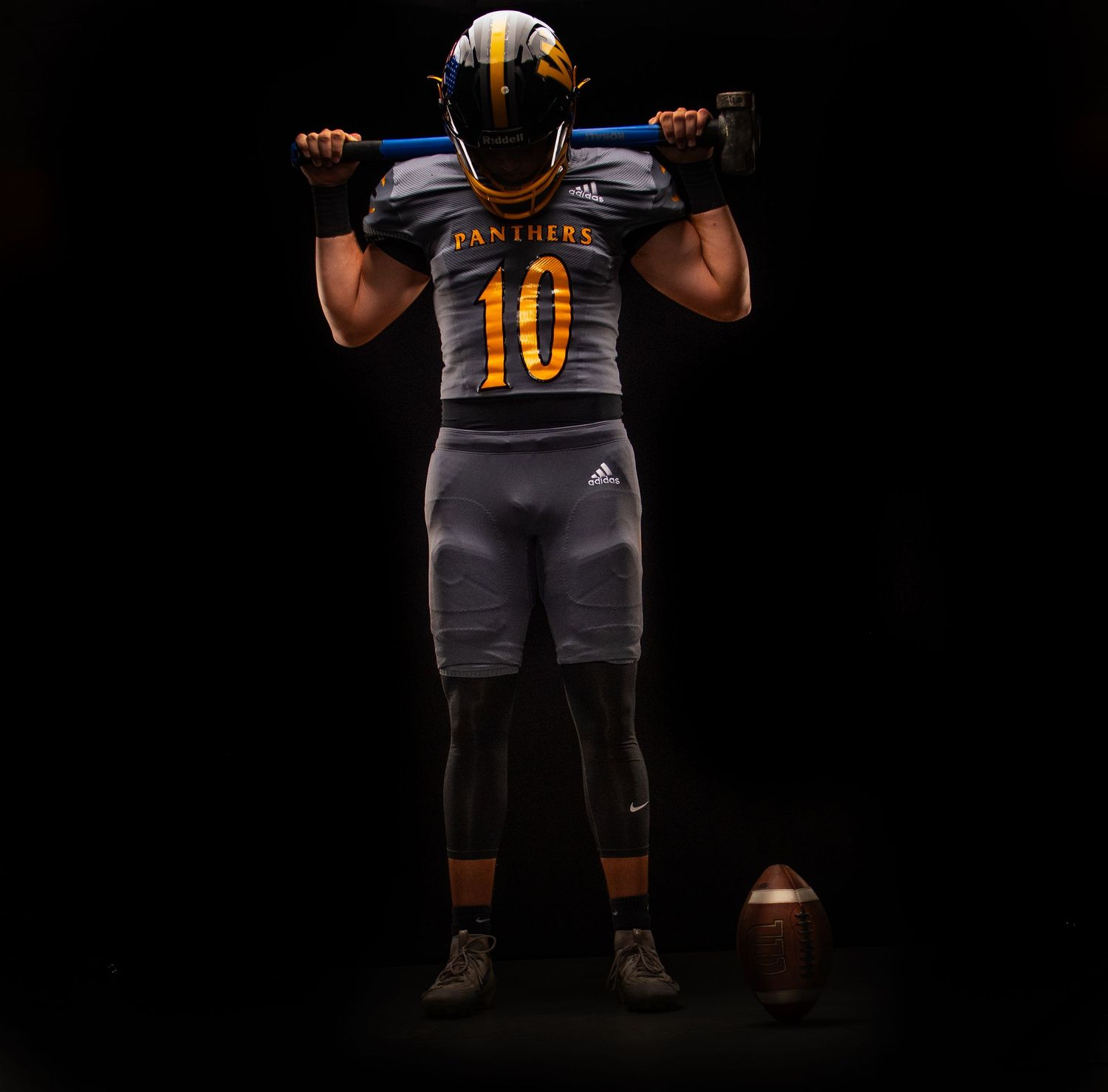 Football player in Panthers #10 gray and gold Adidas uniform holding a bar across shoulders with a football at their feet.