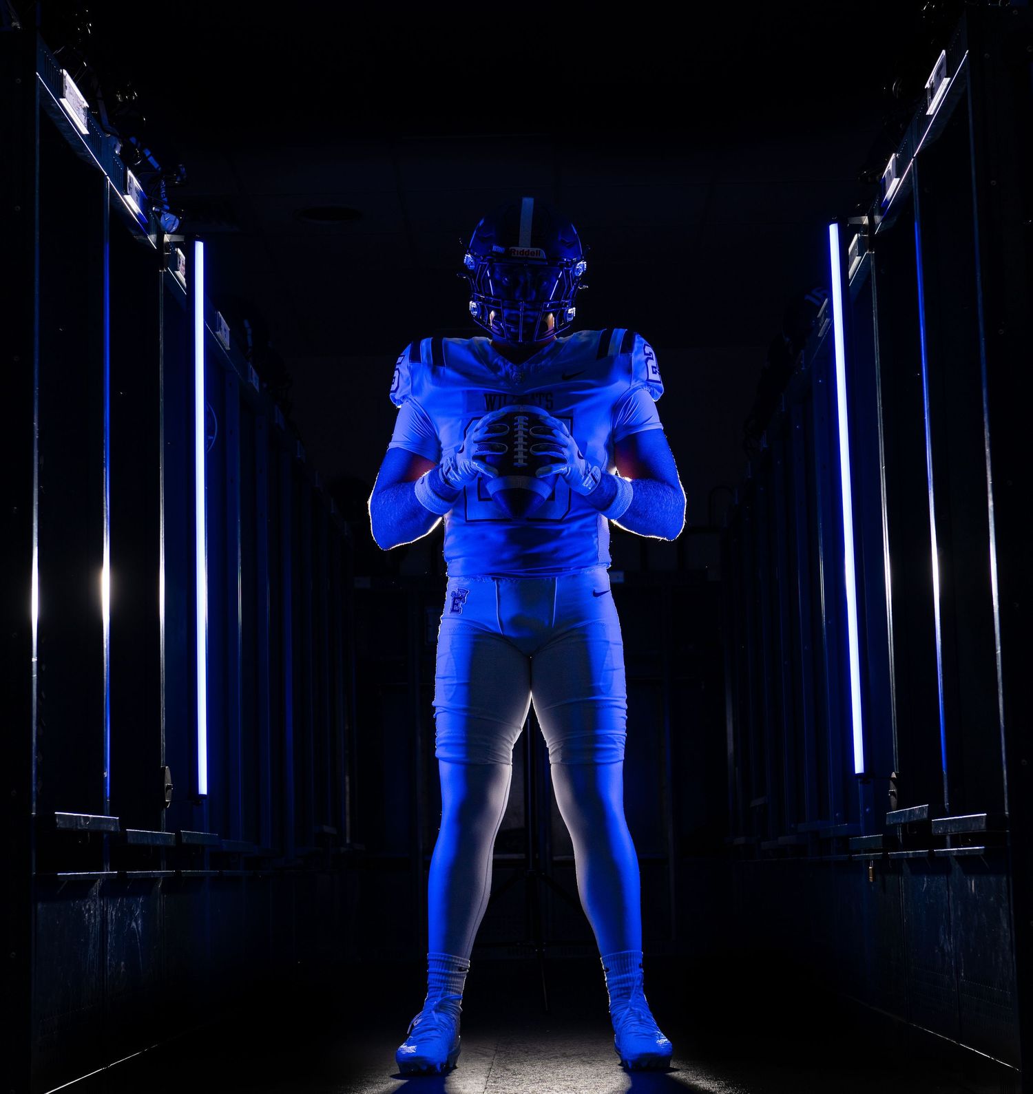 Football player in full blue uniform and helmet stands in a dramatic blue-lit tunnel holding a football.