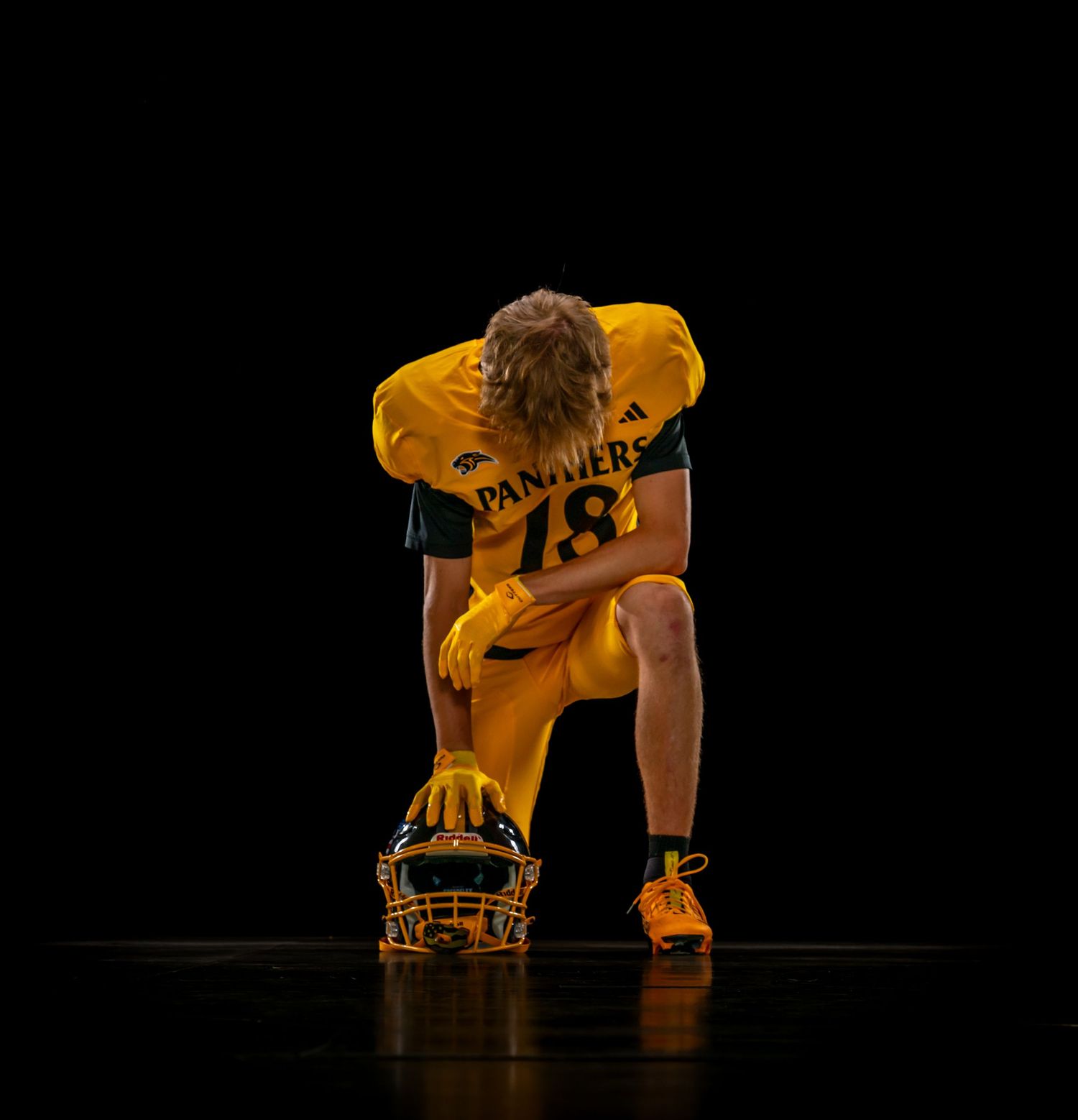 Football player #18 kneels on one knee with helmet on ground in dramatic dark studio shot.