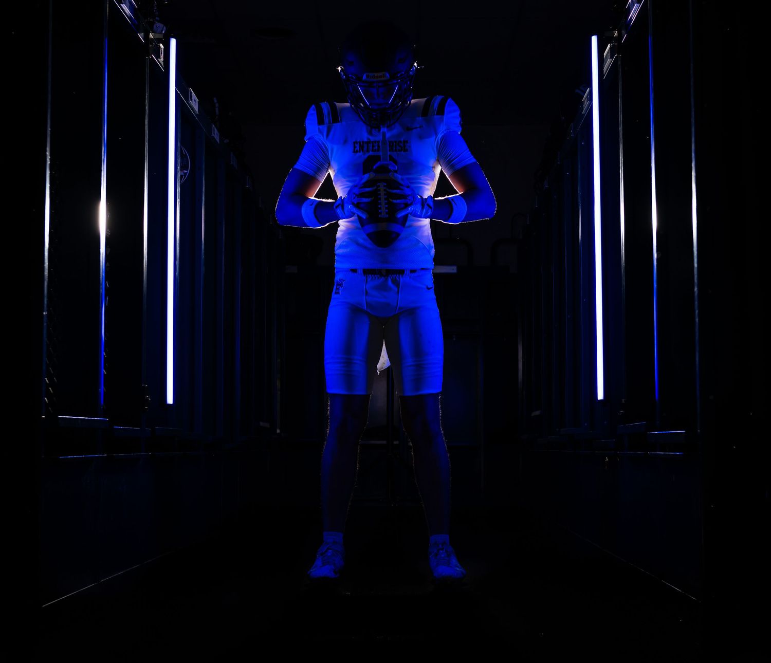 Football player in full gear stands in dark tunnel with blue neon lighting.