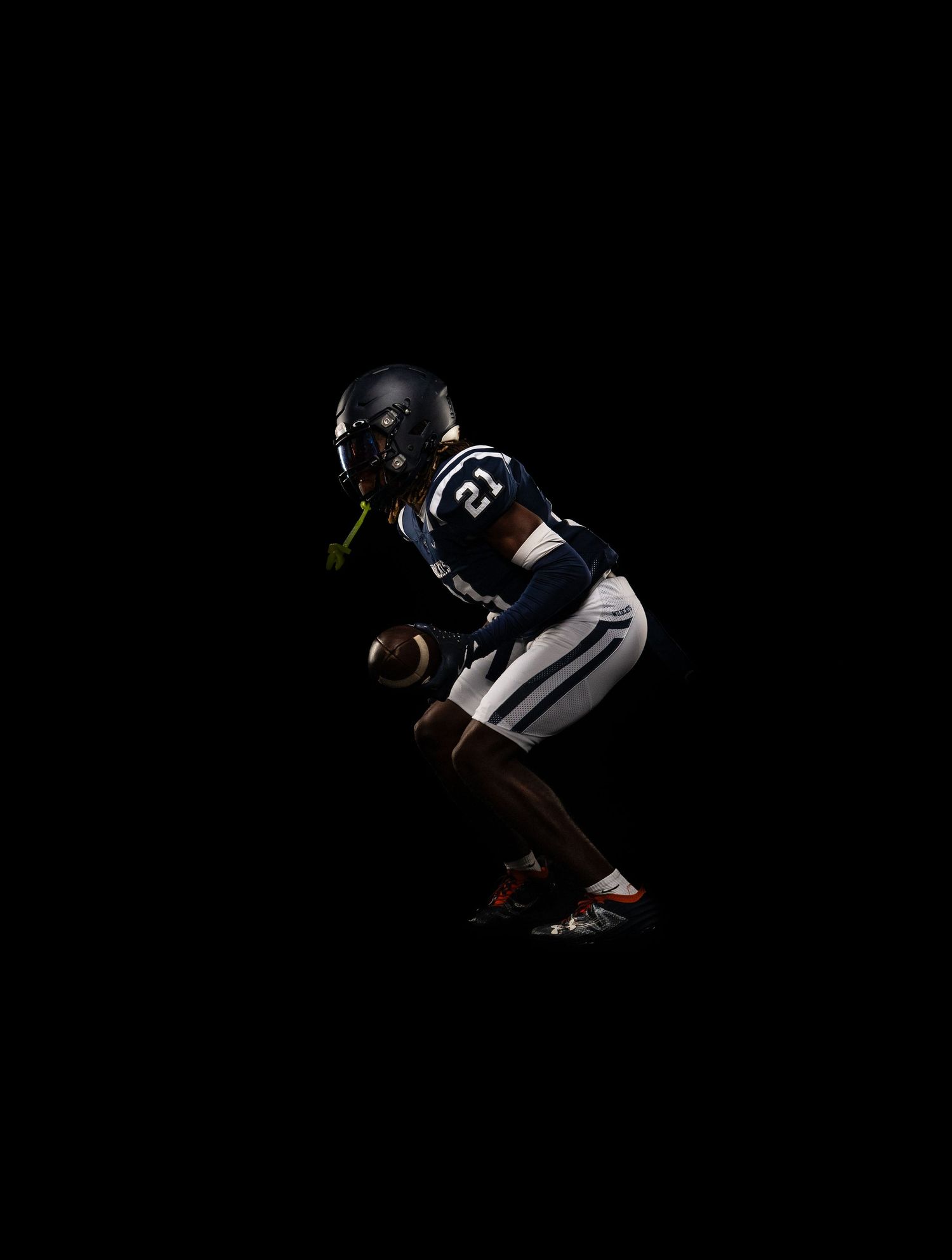 Football player in navy uniform and helmet crouches in ready position holding ball against dark background.